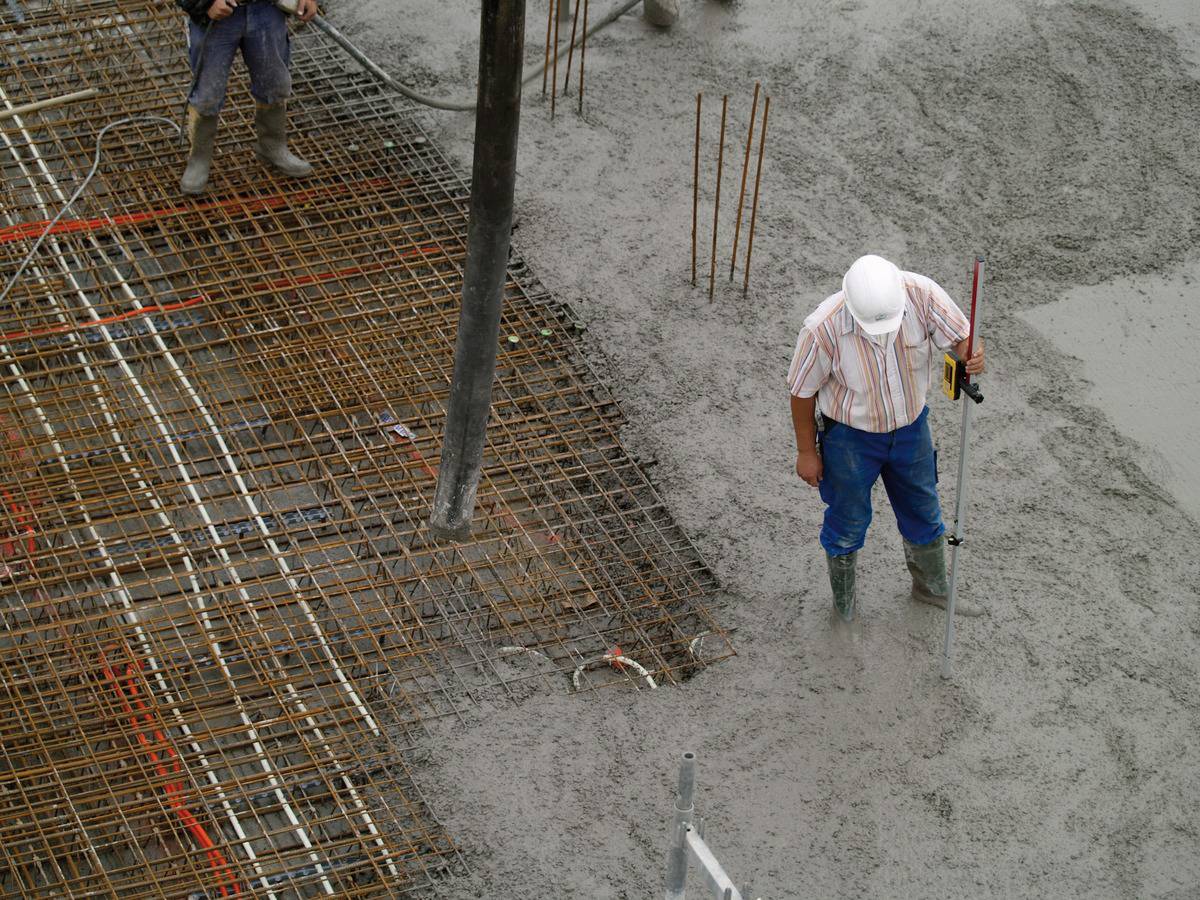 Ein Bauarbeiter mit Helm hält einen Messstab und steht auf einer Baustelle, umgeben von nassem Beton und Stahlverstrebungen.