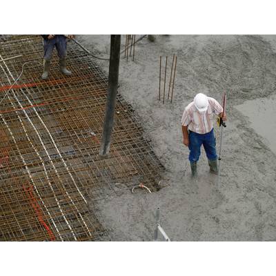 Ein Bauarbeiter mit Helm hält einen Messstab und steht auf einer Baustelle, umgeben von nassem Beton und Stahlverstrebungen.