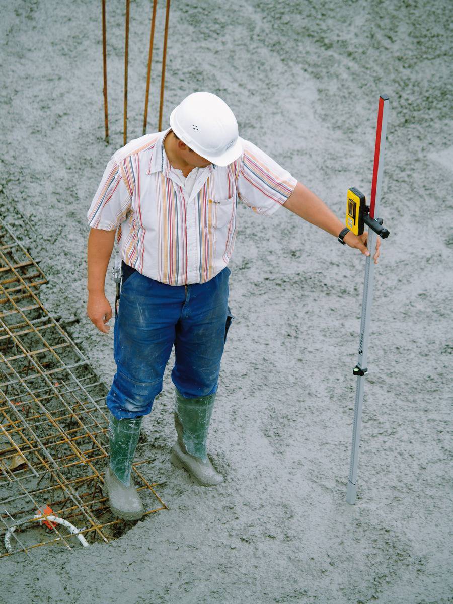 Ein Bauarbeiter in Schutzkleidung steht auf einer Betonfläche und hält ein Vermessungsgerät, um die Betonhöhe zu überprüfen.