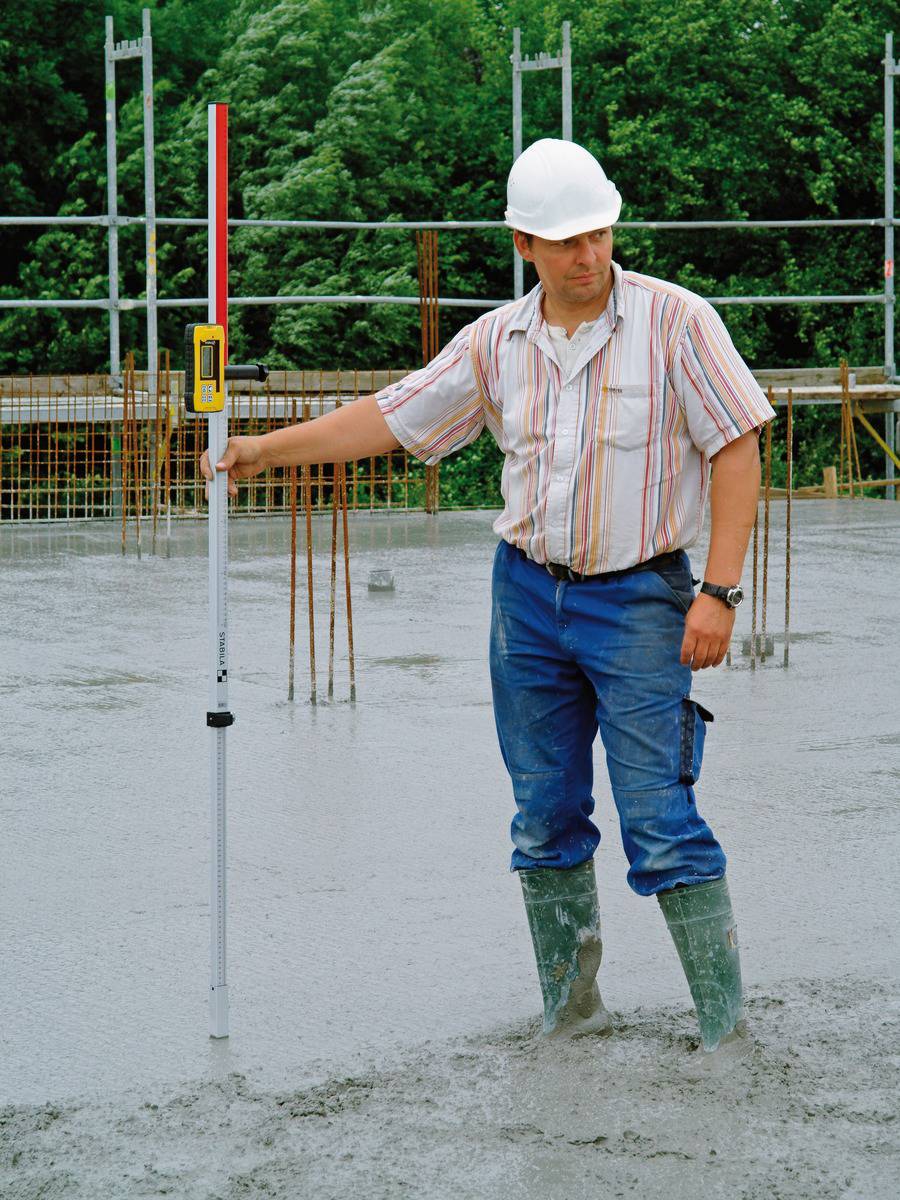 Ein Bauarbeiter mit Schutzhelm misst die Betondecke mit einer Messlatte auf einer Baustelle. Hintergrund: grüne Bäume und Gerüst.