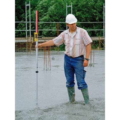 Ein Bauarbeiter mit Schutzhelm misst die Betondecke mit einer Messlatte auf einer Baustelle. Hintergrund: grüne Bäume und Gerüst.