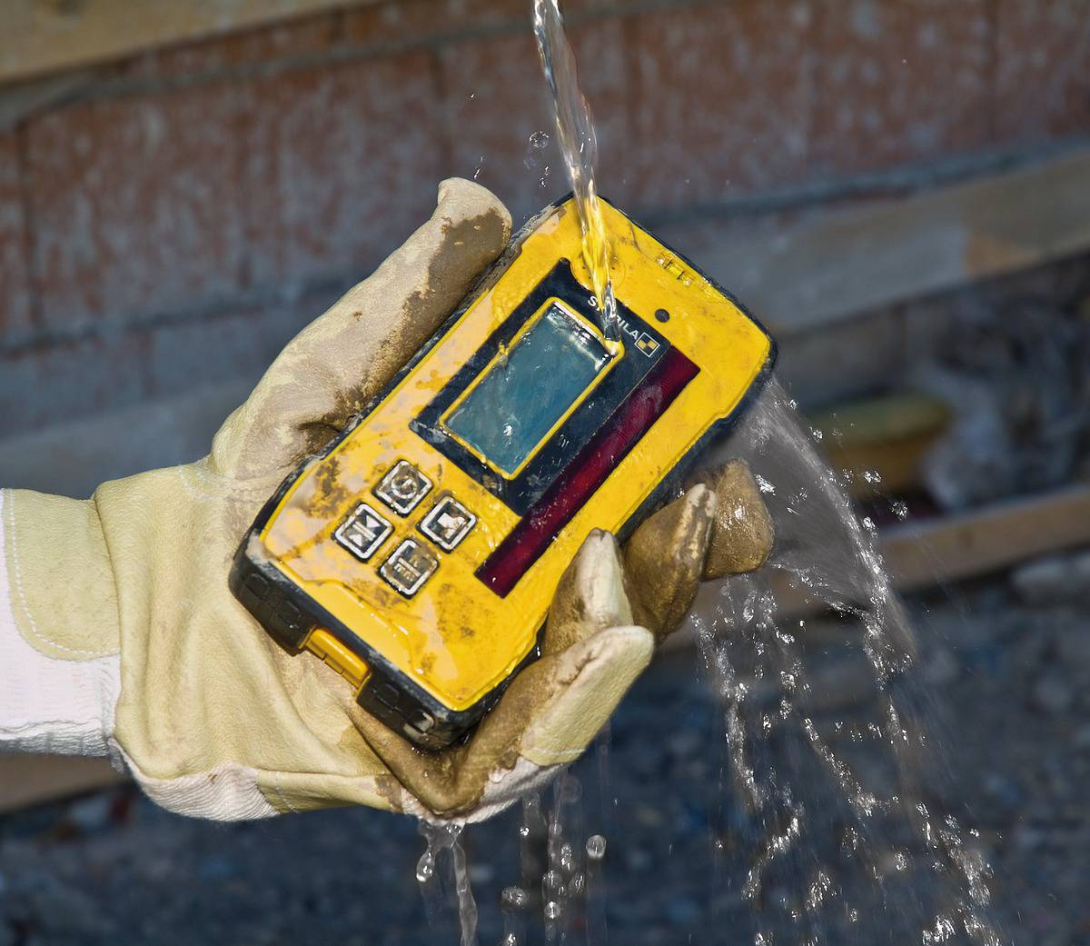 Eine behandschuhte Hand hält ein wasserdichtes, gelbes elektronisches Gerät, das von einem laufenden Wasserstrahl getroffen wird.
