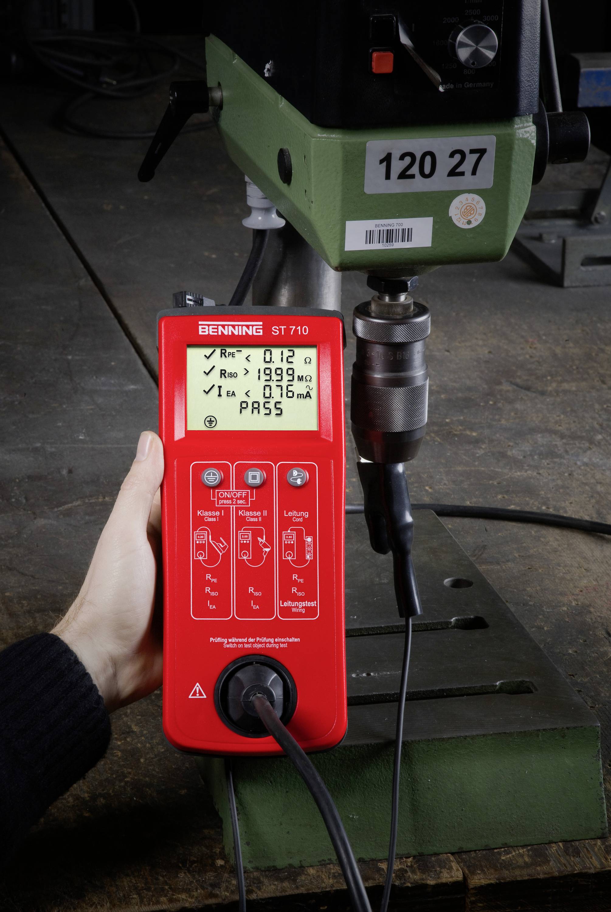 Eine Person hält ein rotes Prüfgerät, das elektrische Messungen an einer grünen Maschine durchführt. Der Bildschirm zeigt Messwerte an.