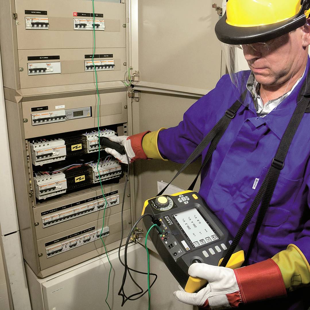 Elektriker mit Schutzhelm und Handschuhen überprüft eine elektrische Schalttafel mit einem Prüfgerät in einem Technikraum.