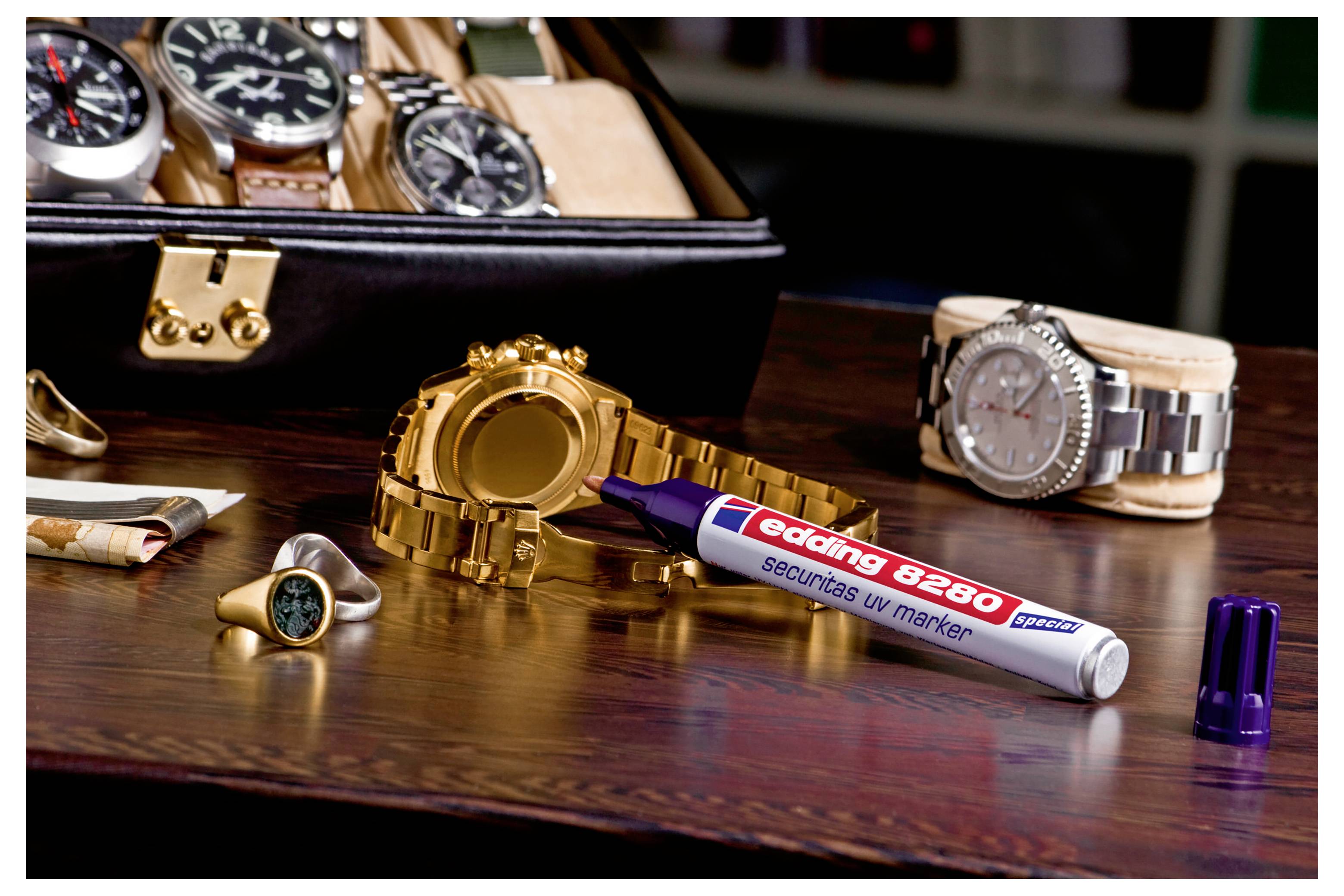 Goldene Armbanduhr, Ring und silberne Uhr auf einem Holztisch, mit einem 'edding 8280' Sicherheitsmarkerstift im Vordergrund.
