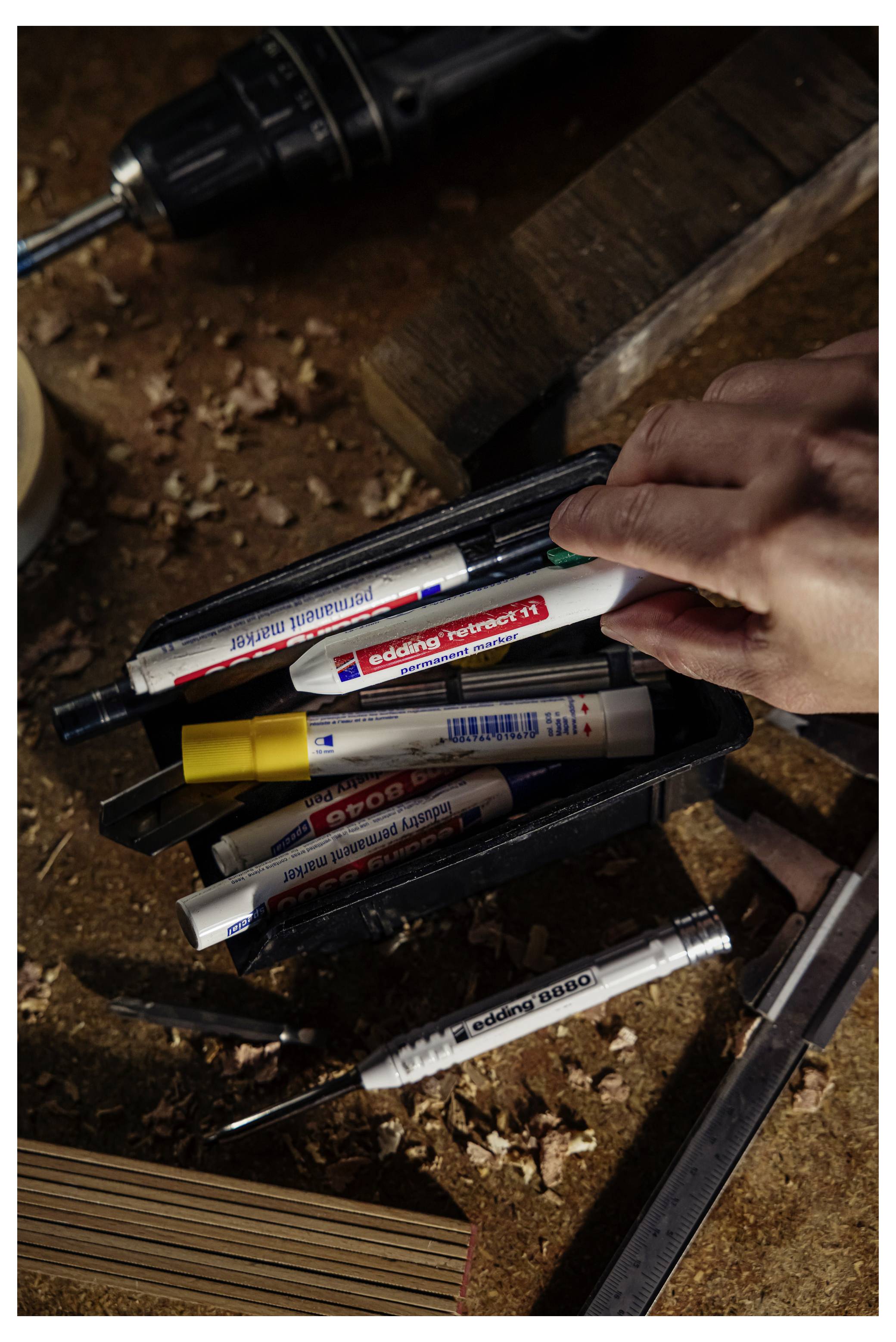 Eine Hand nimmt einen schwarzen Marker aus einer Schachtel mit verschiedenfarbigen Permanentmarkern von einer staubigen Werkbank auf, neben der sich ein Bohrmaschine befindet.