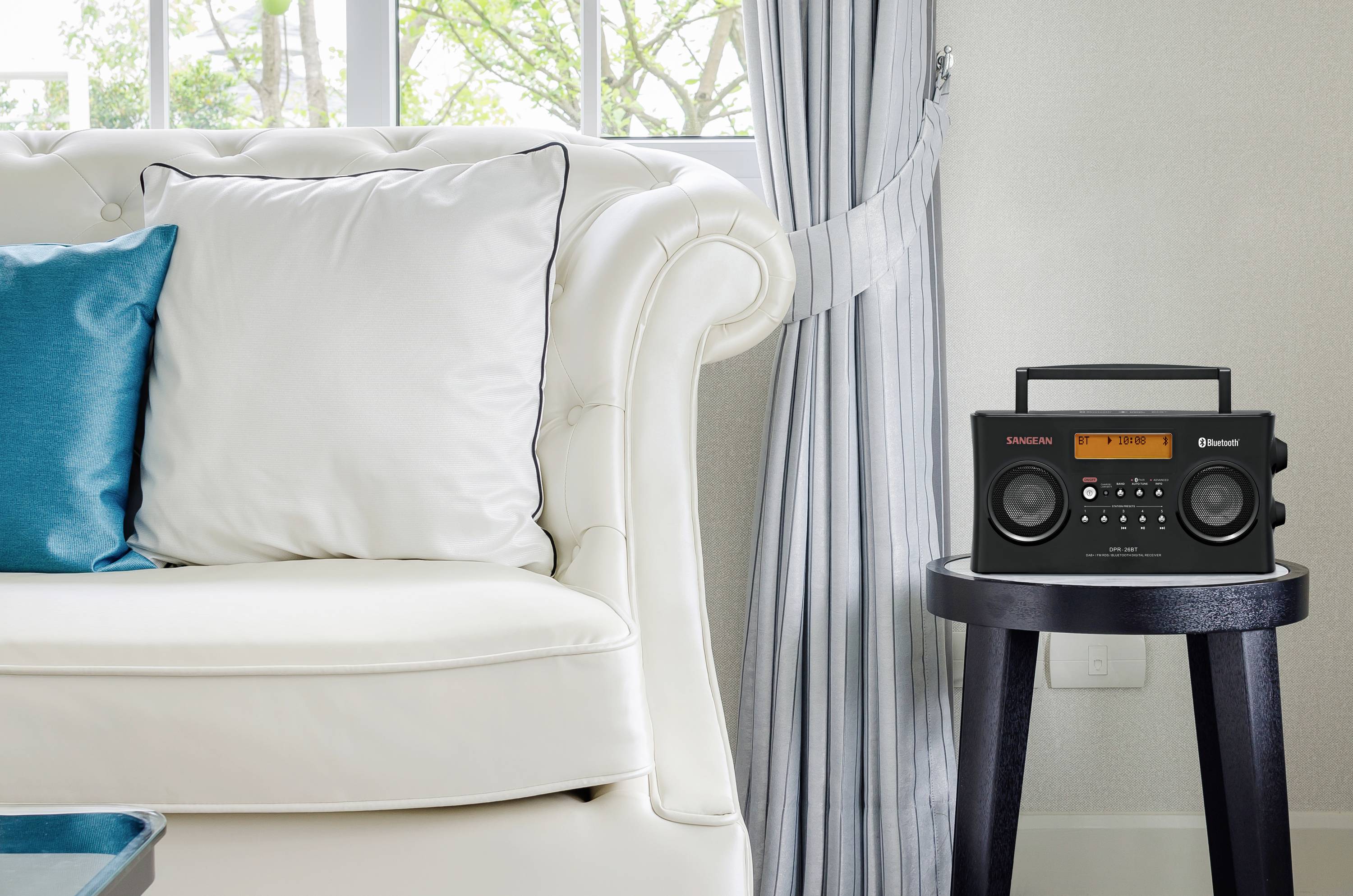 Ein elegantes Wohnzimmer mit weißem Sofa und blauem Kissen. Rechts steht auf einem kleinen Tisch ein schwarzes Radio mit Display.