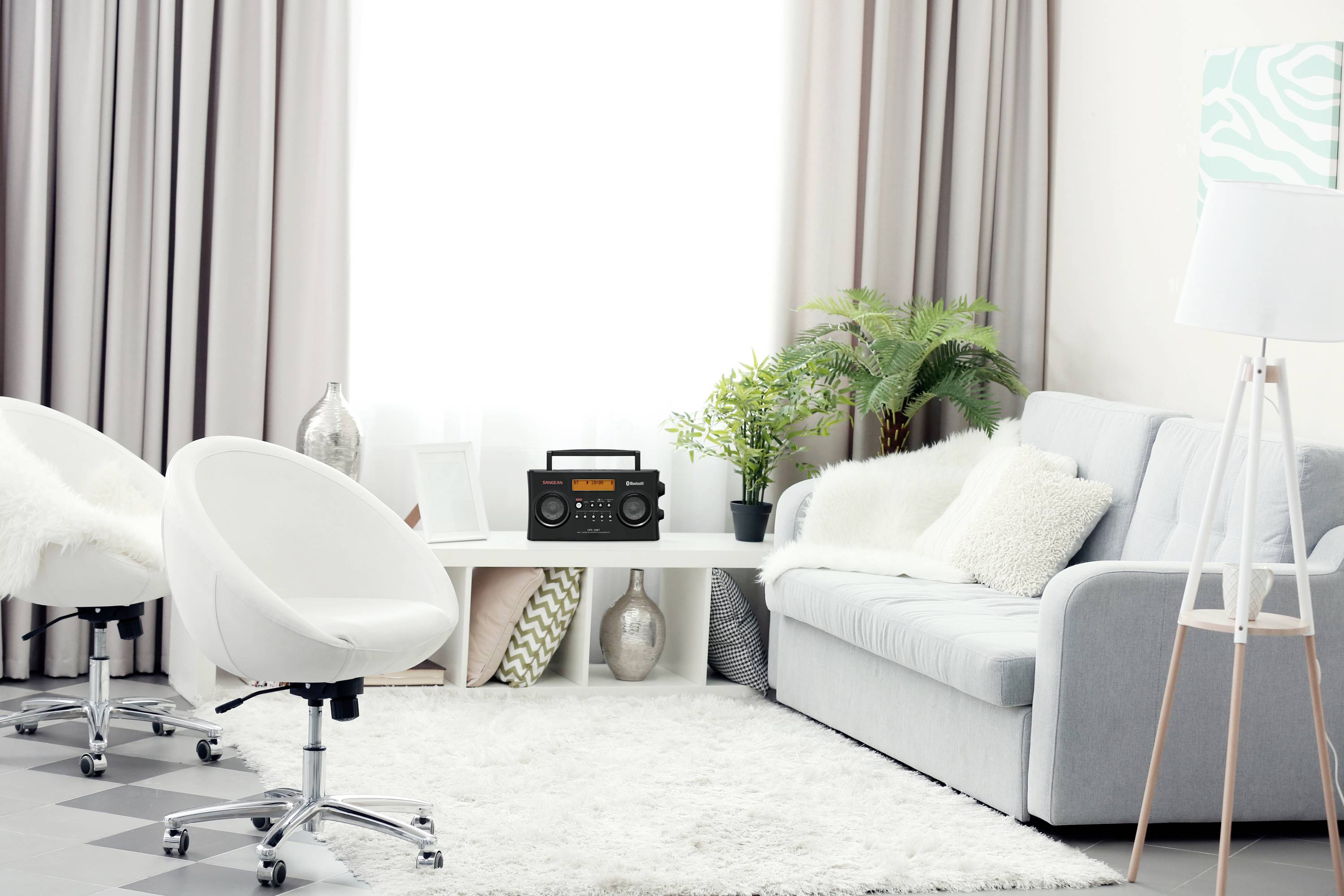 Helles Wohnzimmer mit grauem Sofa, zwei weißen Stühlen, einem Couchtisch mit Radio und Pflanzen vor einem großen Fenster mit Vorhängen.