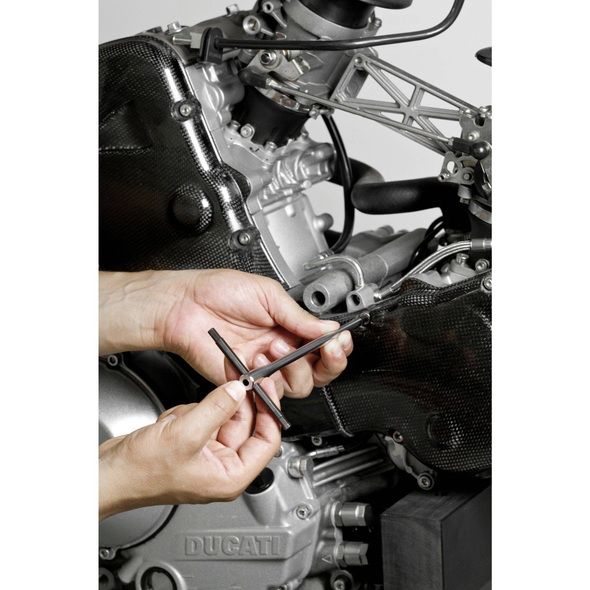 Eine Person hält ein T-Schlüsselwerkzeug und arbeitet an einem Motorradmotor mit sichtbaren Teilen aus Metall und Karbon.