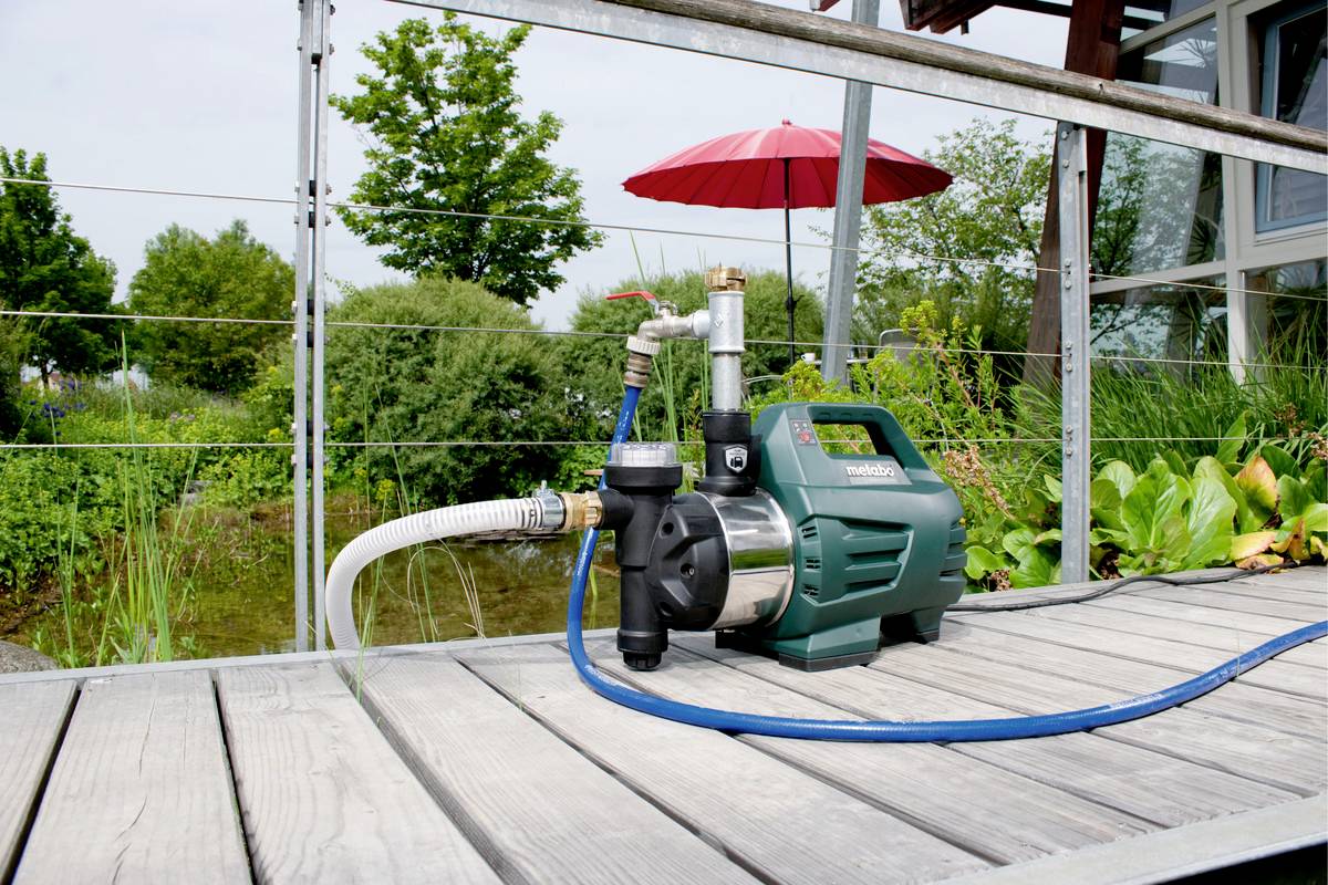Wasserpumpe steht auf einer Holzterrasse im Garten mit Pflanzen, einem roten Sonnenschirm und einem modernen Gebäude im Hintergrund.