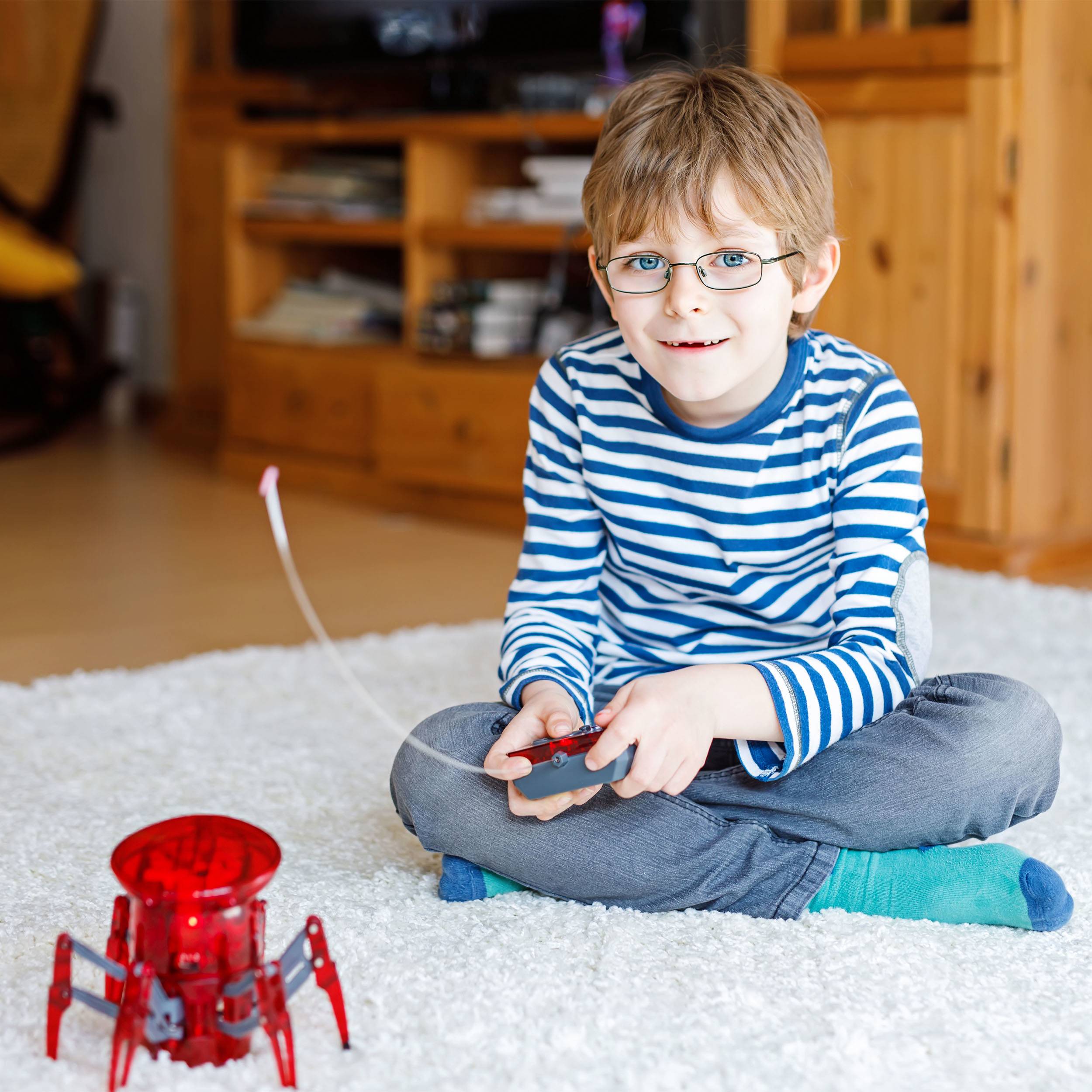 Ein Junge sitzt auf einem Teppich und steuert mit einem Controller einen roten Spielzeugroboter. Im Hintergrund Möbel.