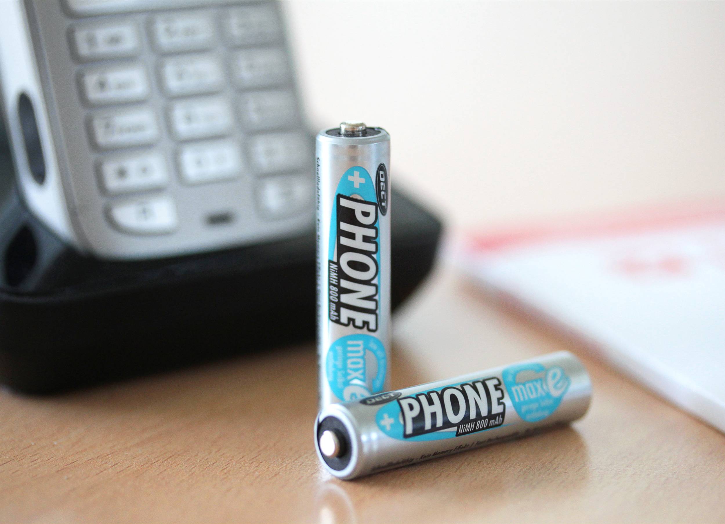 Zwei Batterien der Marke 'Phone' stehen auf einem Tisch vor einem Telefon mit sichtbarem Tastenfeld.