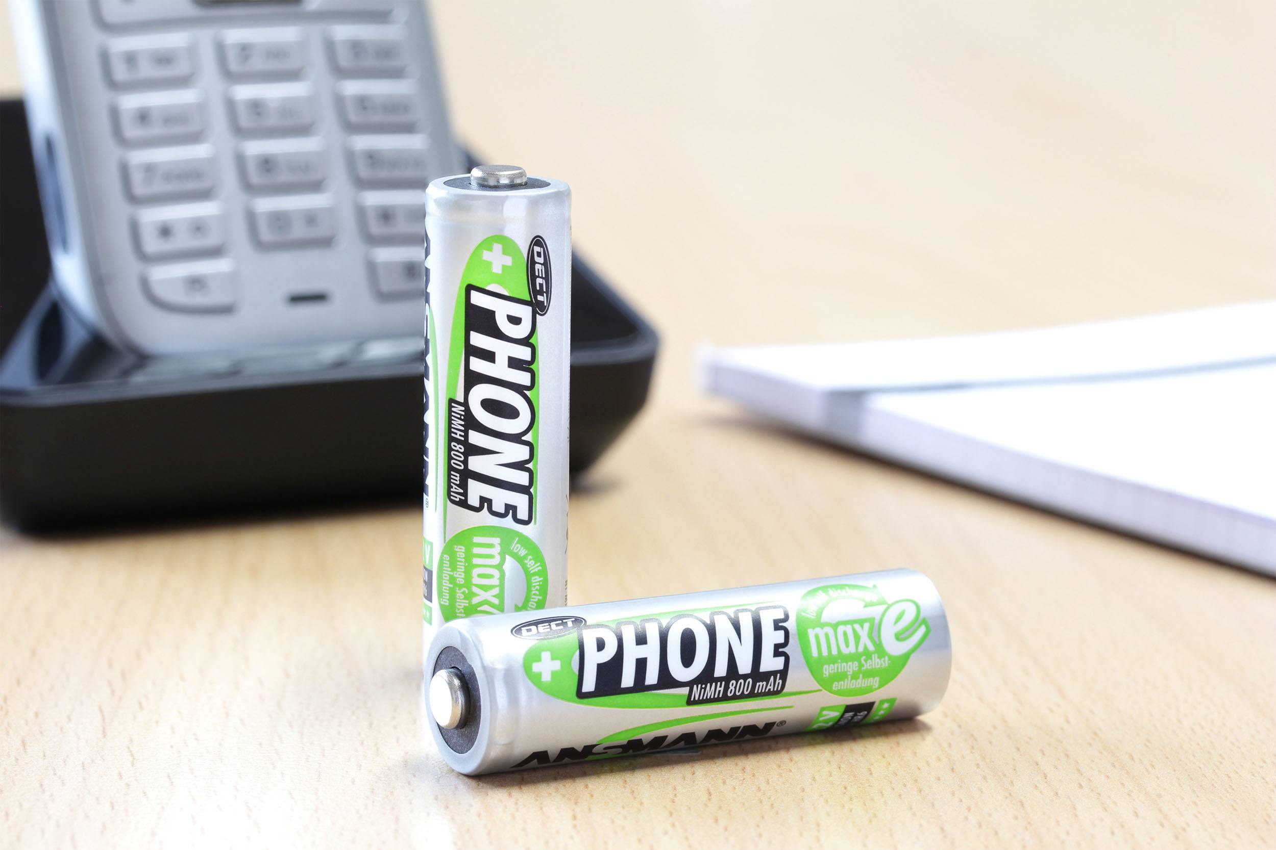 Zwei grüne Telefonbatterien auf einem Schreibtisch vor einem kabellosen Telefon. Eine der Batterien steht, die andere liegt davor.