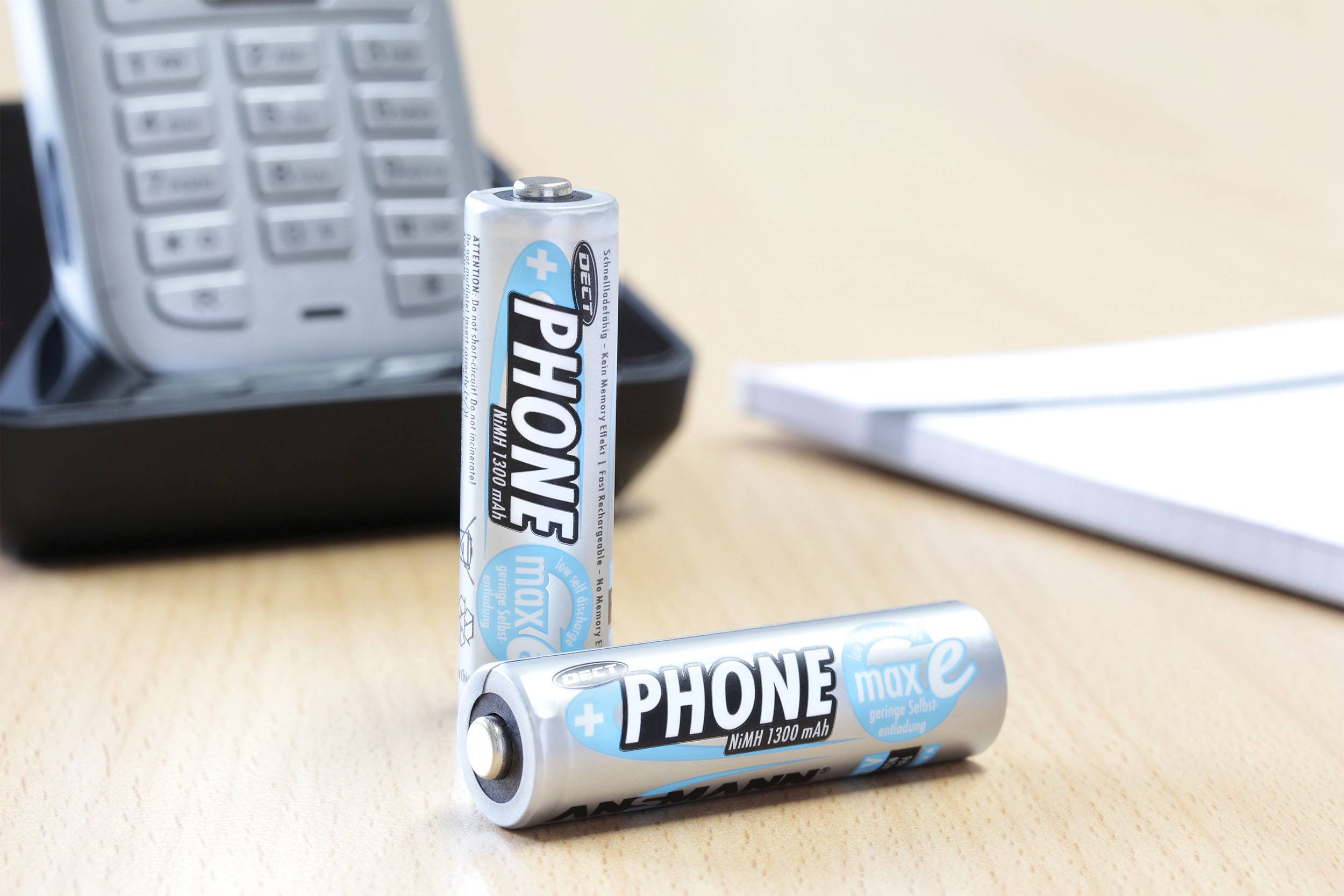 Zwei AA-Batterien mit der Aufschrift 'PHONE', eine aufrecht, die andere liegend, vor einem schnurlosen Telefon auf einem Schreibtisch.
