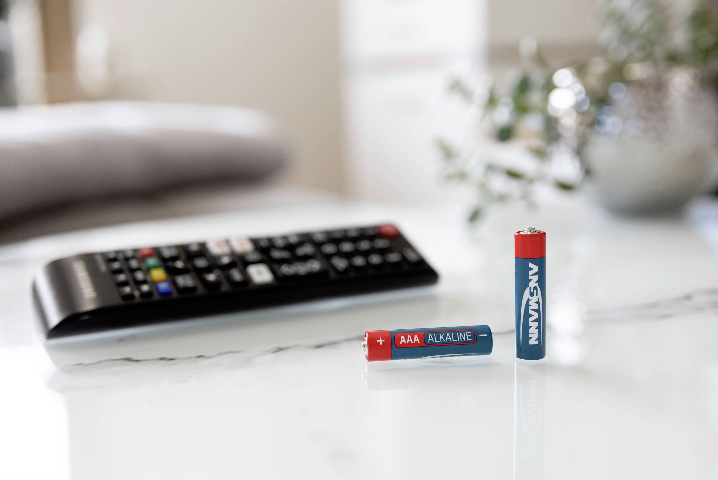 Eine Fernbedienung liegt auf einem Tisch, daneben zwei Batterien, eine liegend ('AAA Alkaline') und eine stehend.