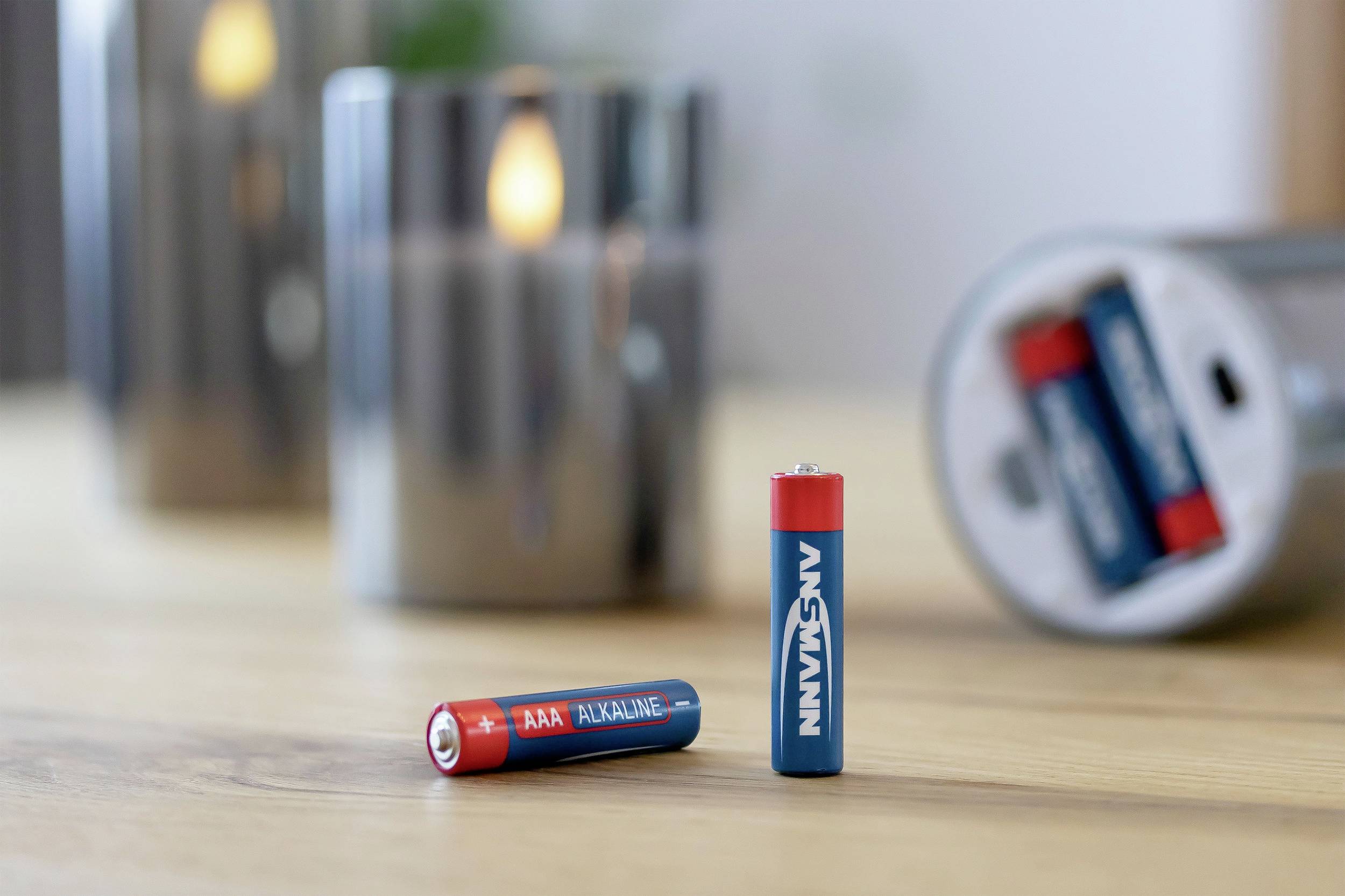 Zwei rote und blaue AAA-Batterien stehen auf einem Tisch; im Hintergrund unscharf eine glänzende Kerze und eine offene Dose mit Batterien.