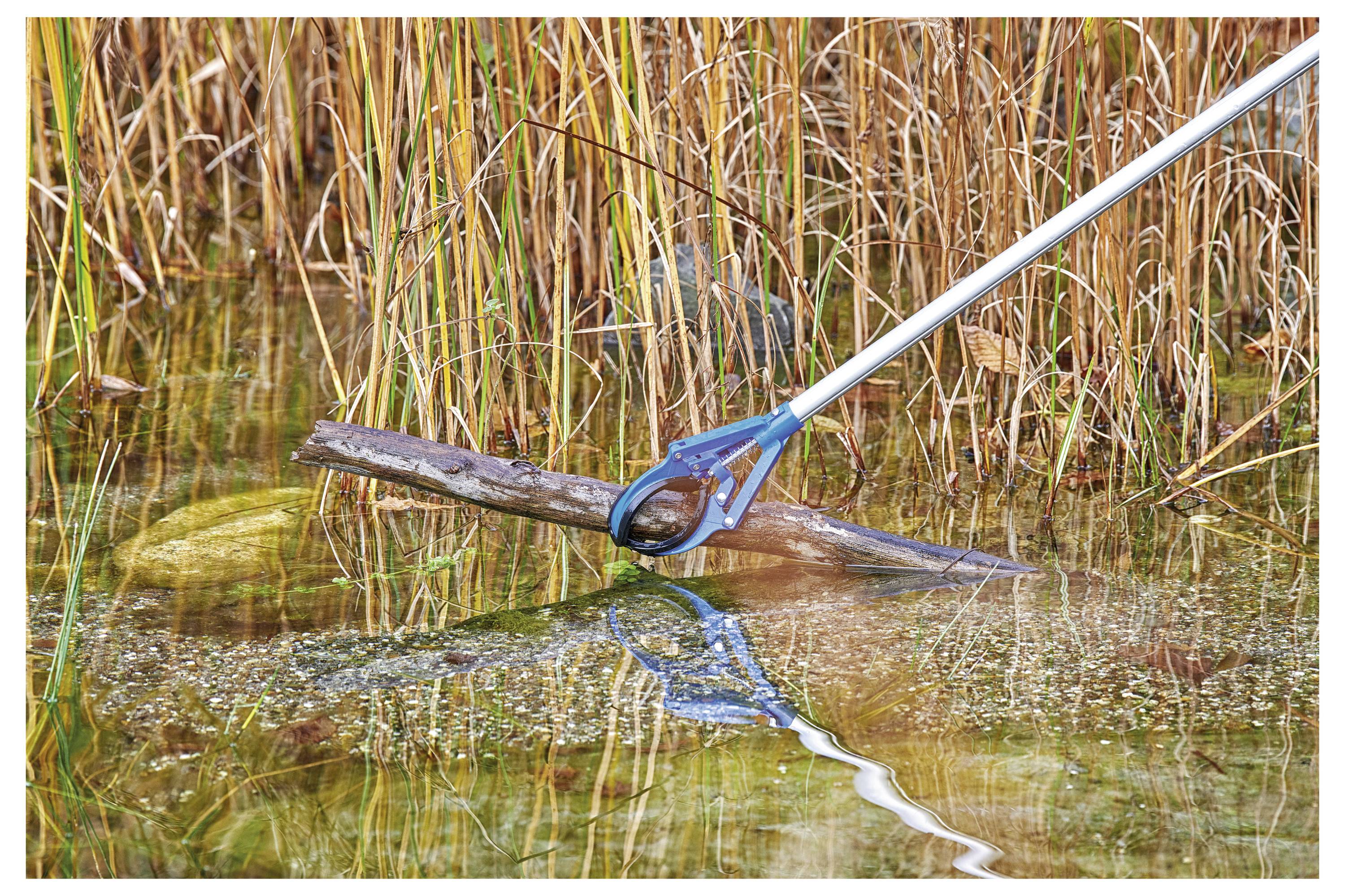 Eine Person benutzt ein blaues Werkzeug mit verlängertem Griff, um Ablagerungen aus einem flachen, mit Wasser gefüllten Bereich zu entfernen, der von hohen Schilfrohrstauden umgeben ist.