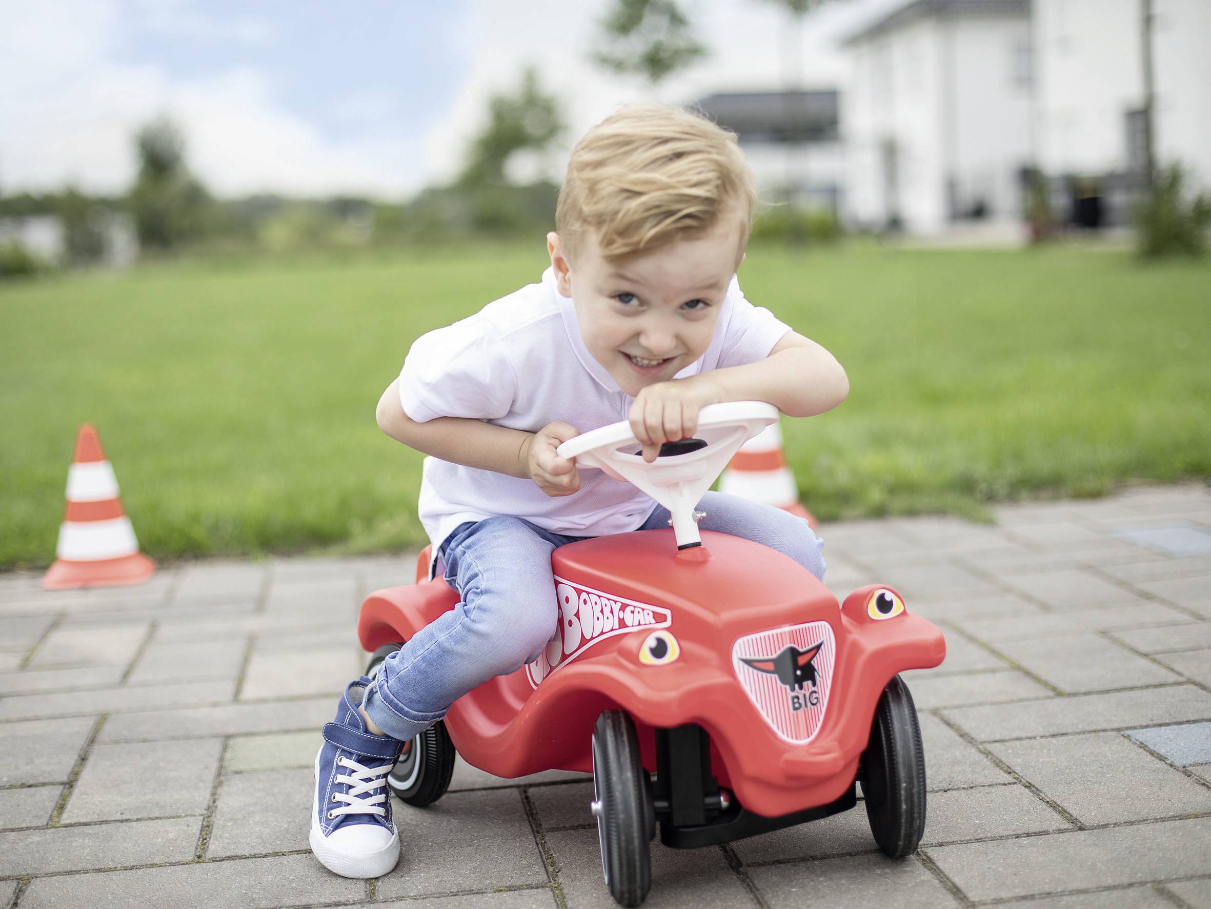 Ein Kind in weißem T-Shirt fährt lachend auf einem roten Spielzeugauto über einen gepflasterten Weg im Freien. Im Hintergrund sind einige Verkehrshütchen und Grünflächen zu sehen.