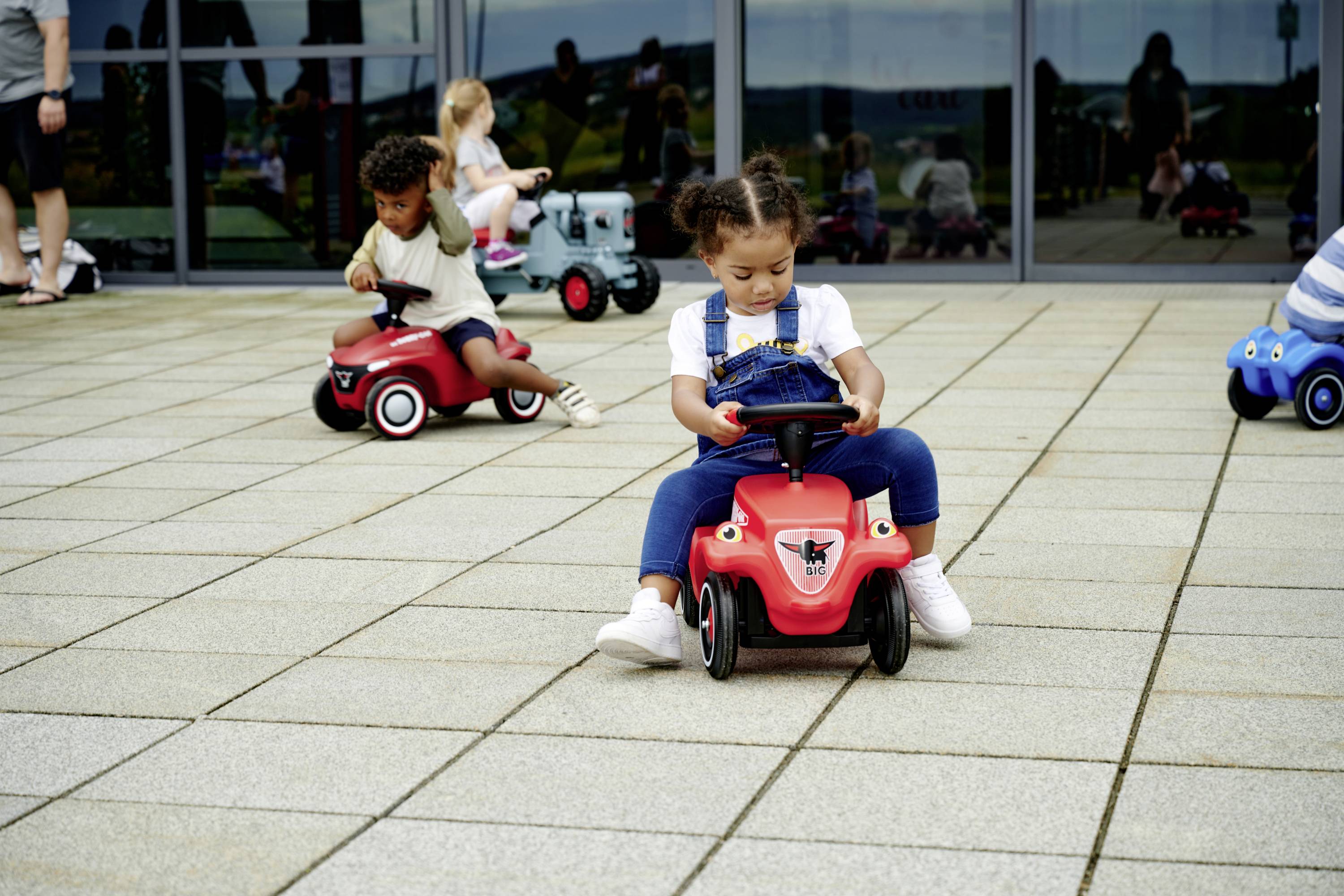 Kinder spielen auf roten Spielzeugautos im Freien vor einem großen Fenstergebäude. Im Hintergrund weitere Kinder und Erwachsene.