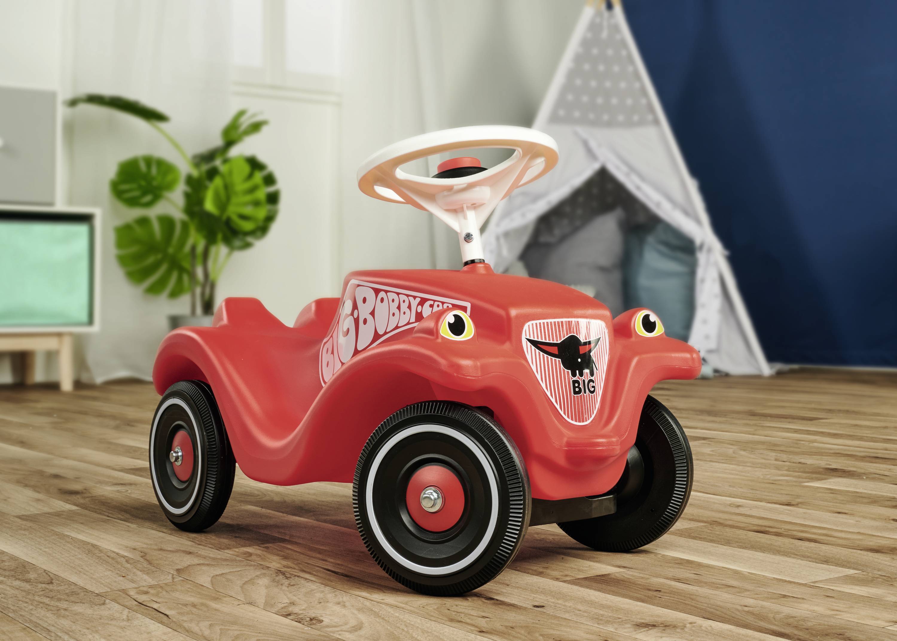 Ein rotes Kinderfahrzeug steht auf einem Holzboden in einem Spielzimmer mit einer Pflanze und einem Zelt im Hintergrund.
