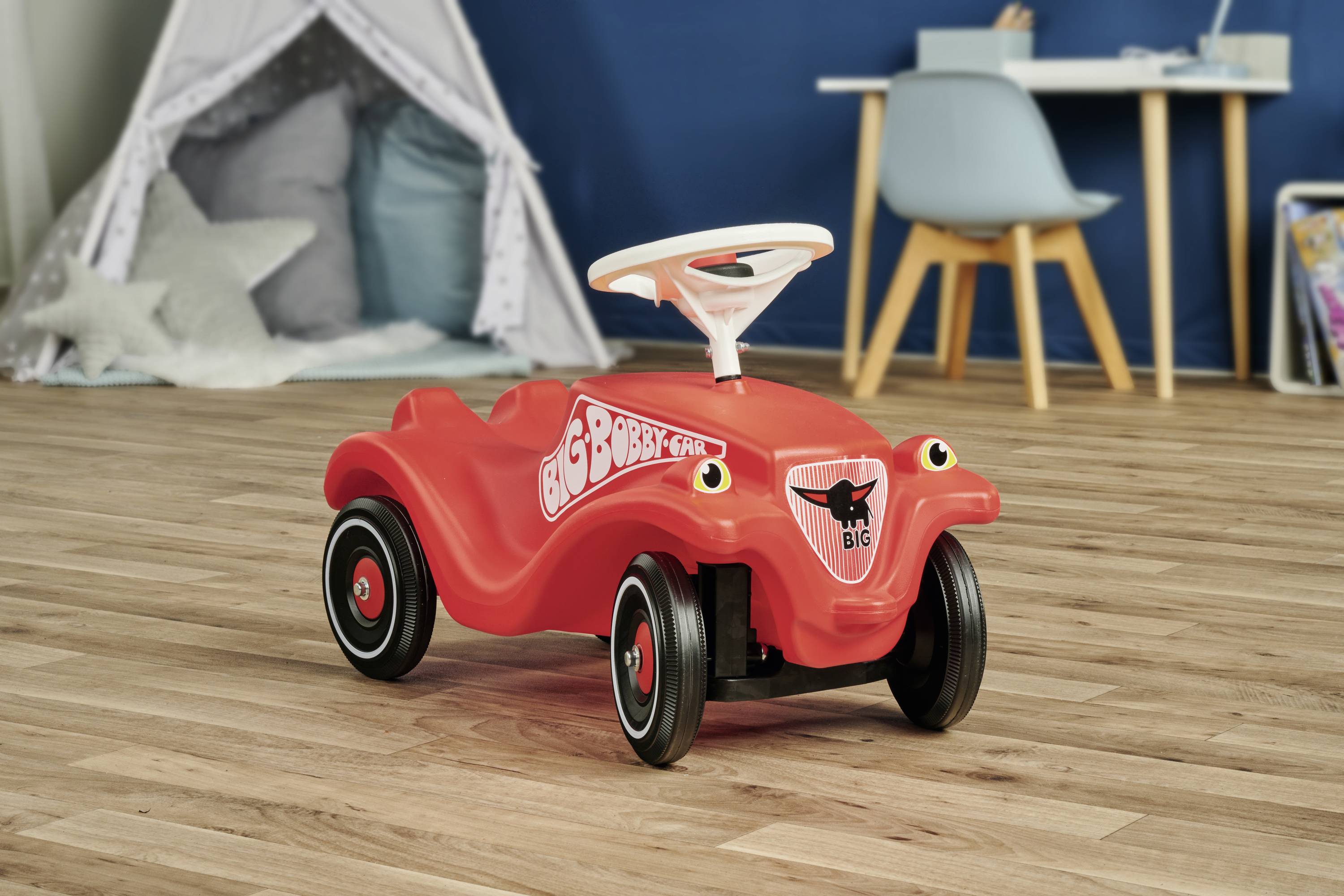 Rotes Kinderspielzeugauto mit der Aufschrift 'BIG Bobby Car', platziert auf Holzfußboden in einem Raum mit blauem Stuhl und Zelt.