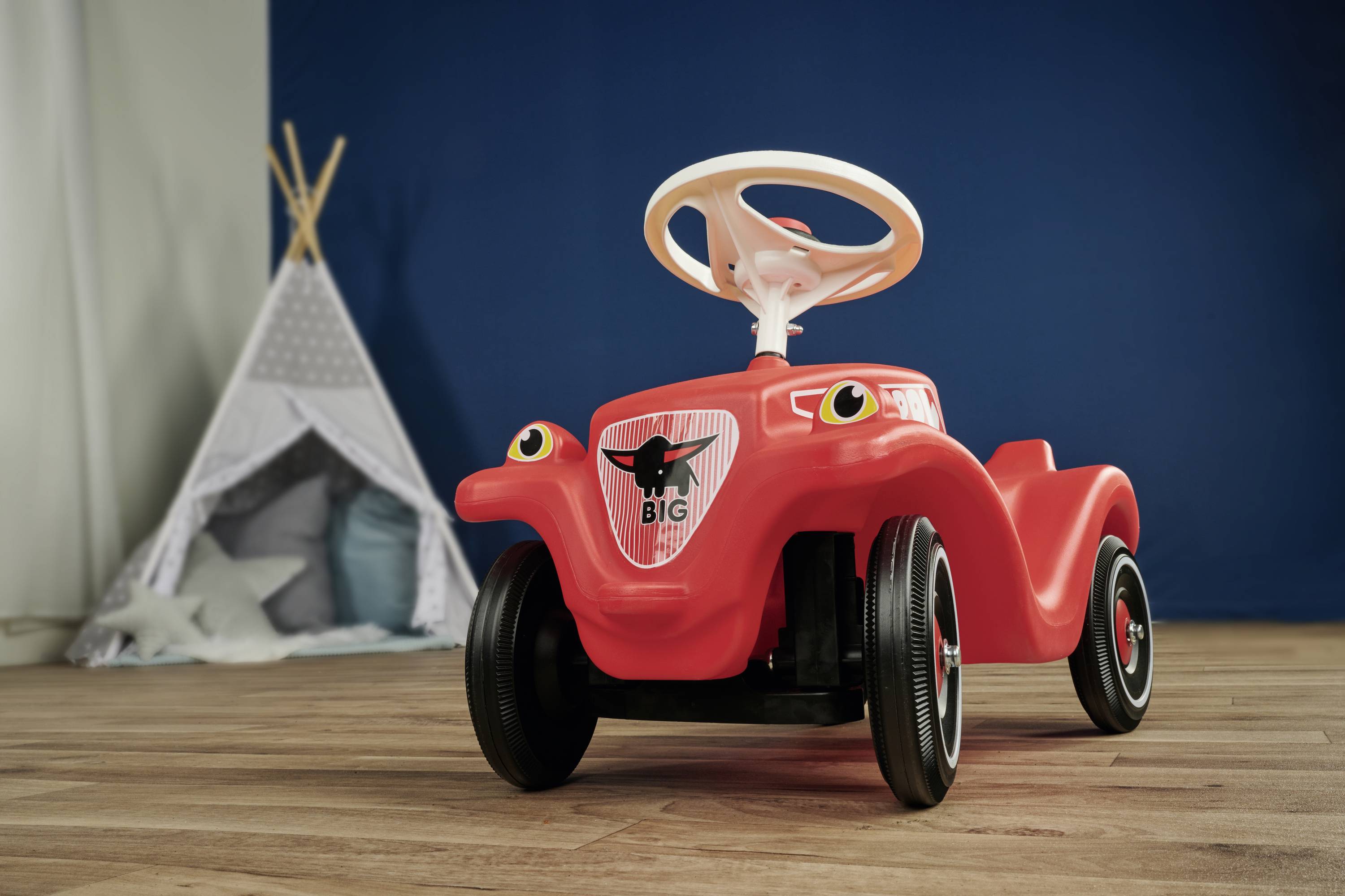 Rotes Spielzeugauto mit weißen Lenkrad auf Holzboden, im Hintergrund ein Kinderzelt.