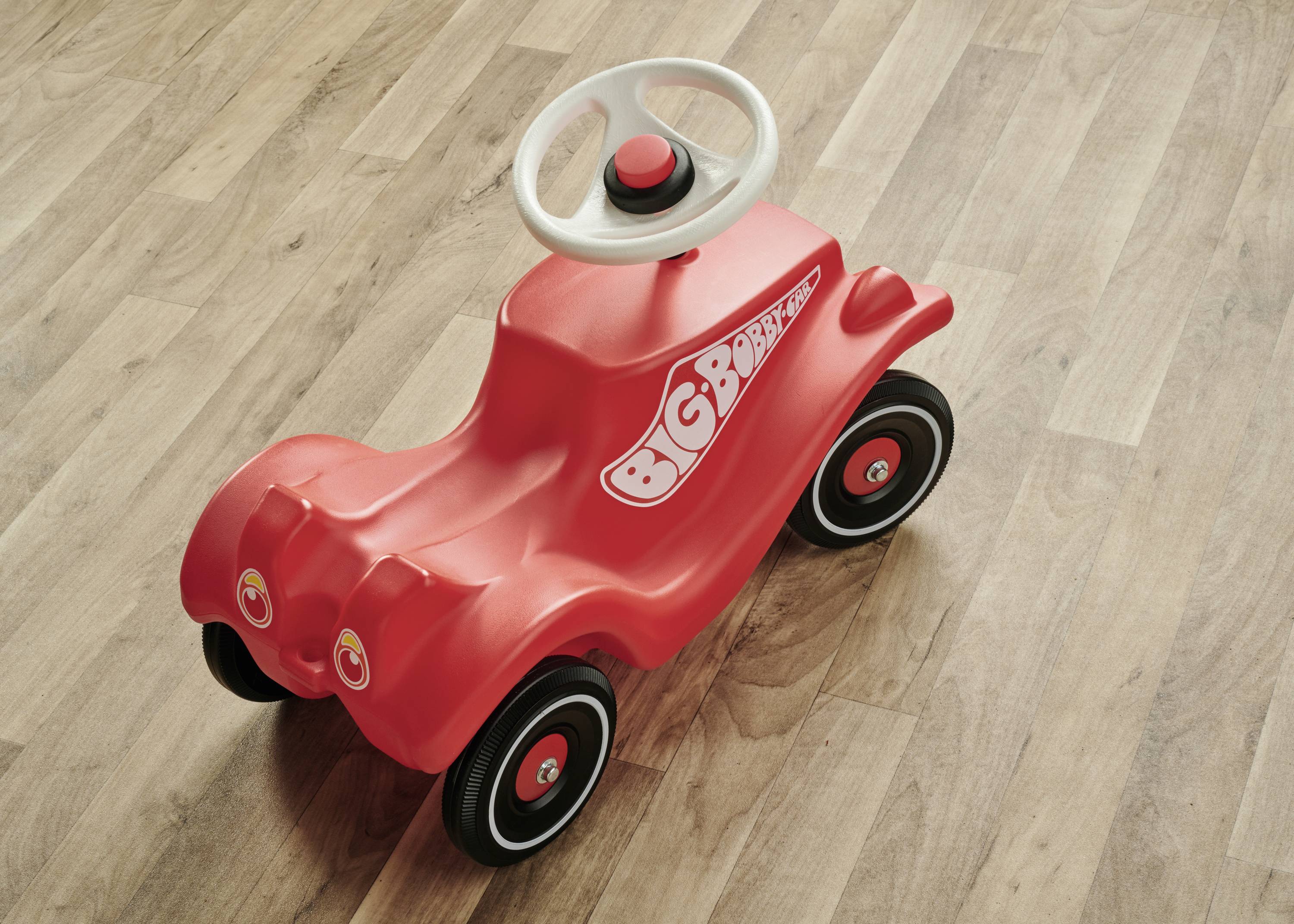 Rotes Kinderfahrzeug mit weißen Lenkrad und schwarzen Rädern, auf Holzboden stehend.