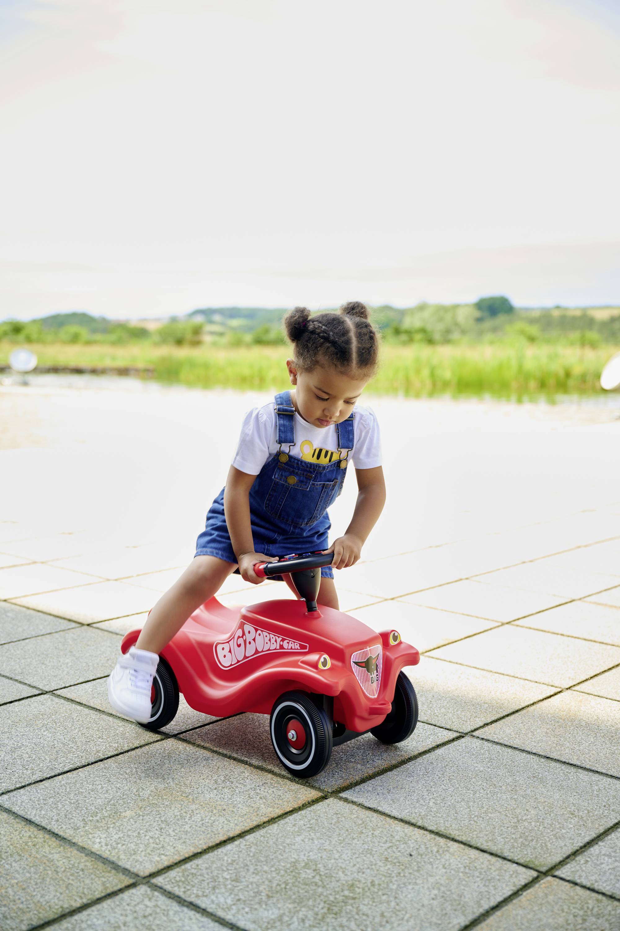 Ein Kind fährt auf einem roten Spielzeugauto im Freien; es trägt einen weißen T-Shirt und blaue Latzhose. Im Hintergrund ist Landschaft erkennbar.