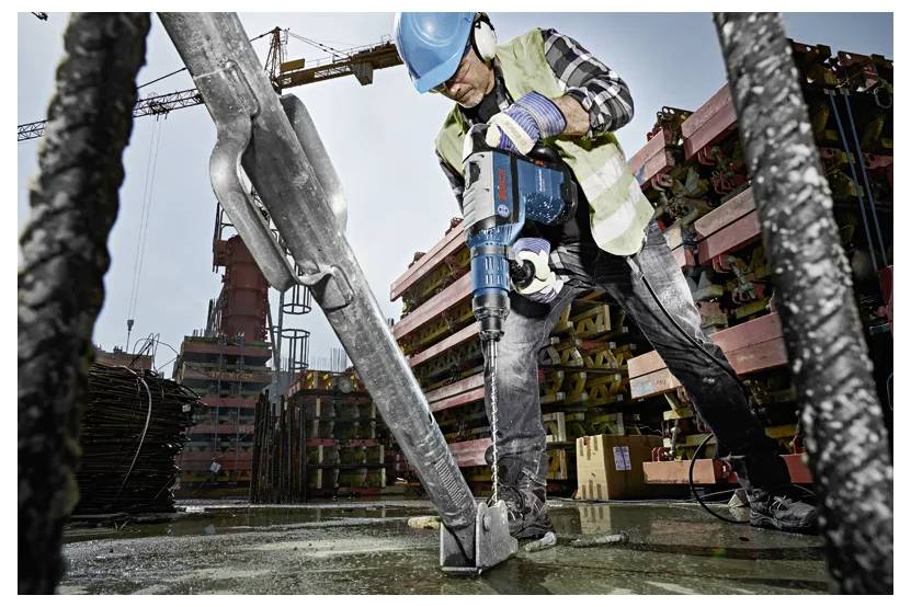 Ein Bauarbeiter mit Sicherheitsausrüstung bedient einen Bohrschrauber auf einer Betonoberfläche auf einer Baustelle, wobei Kräne und Ausrüstung im Hintergrund zu sehen sind.
