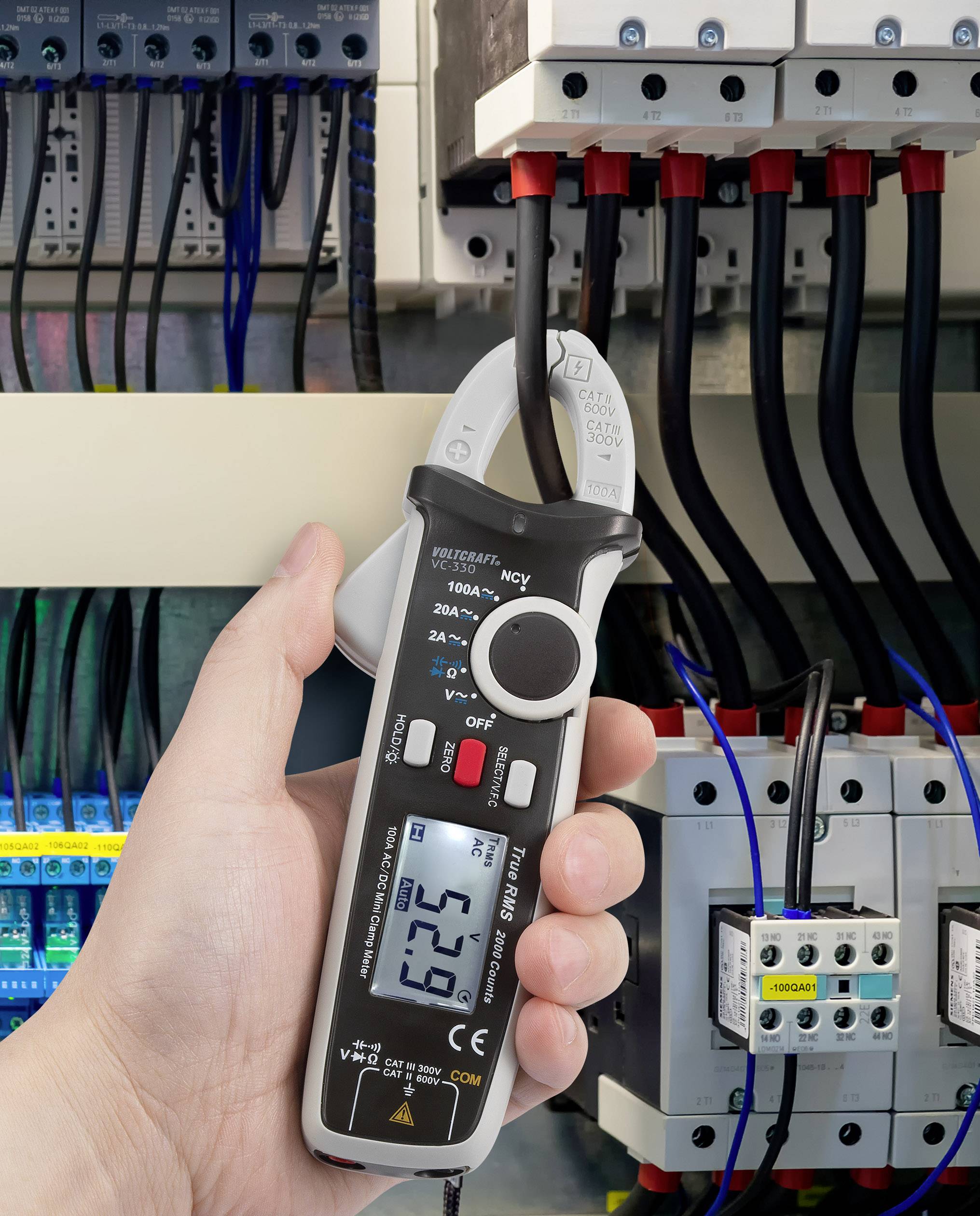 Eine Hand hält ein Digitalmultimeter, das den elektrischen Strom in Kabeln misst, die mit einer Schalttafel verbunden sind.