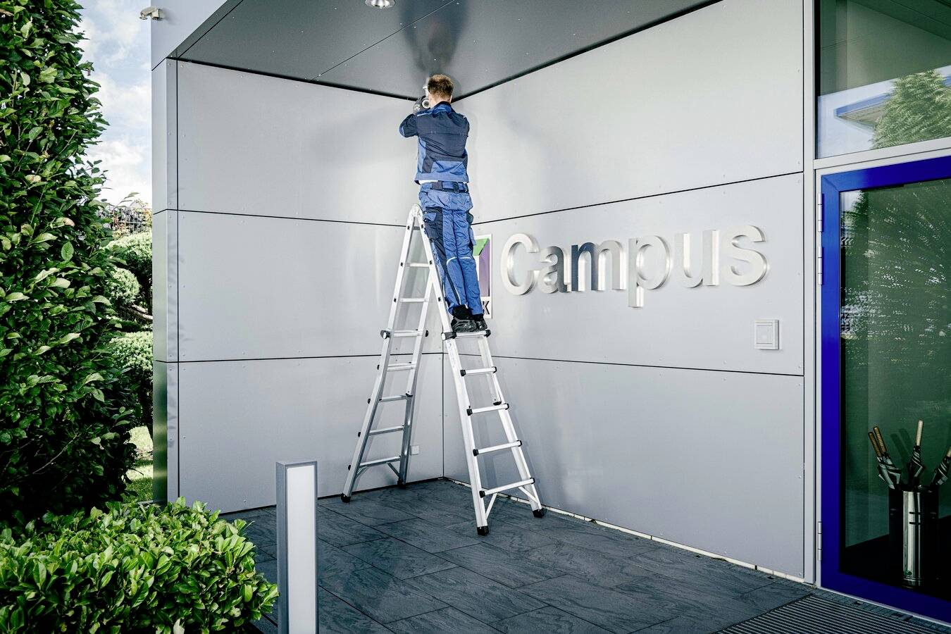 Ein Handwerker auf einer Leiter repariert eine Hauswand vor einem Gebäude mit der Aufschrift 'Campus'.