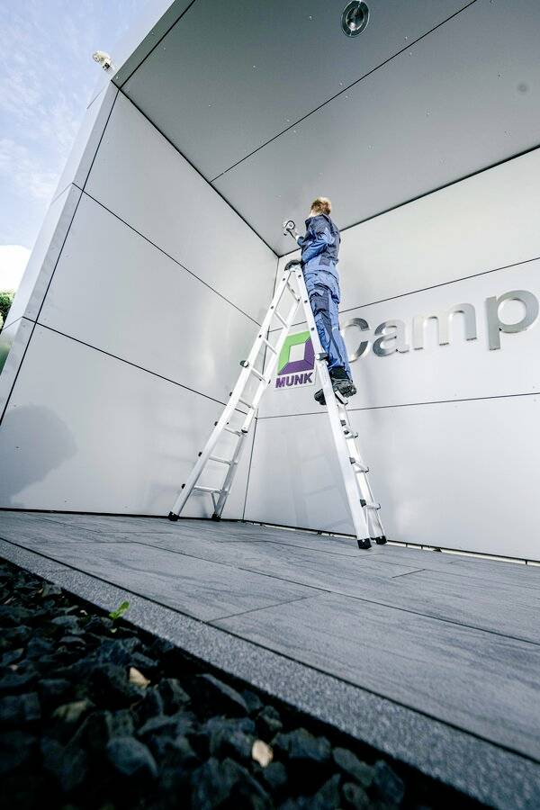 Person auf Leiter arbeitet an Decke eines modernen Gebäudeeingangs. Im Hintergrund ist ein Firmenlogo sichtbar.