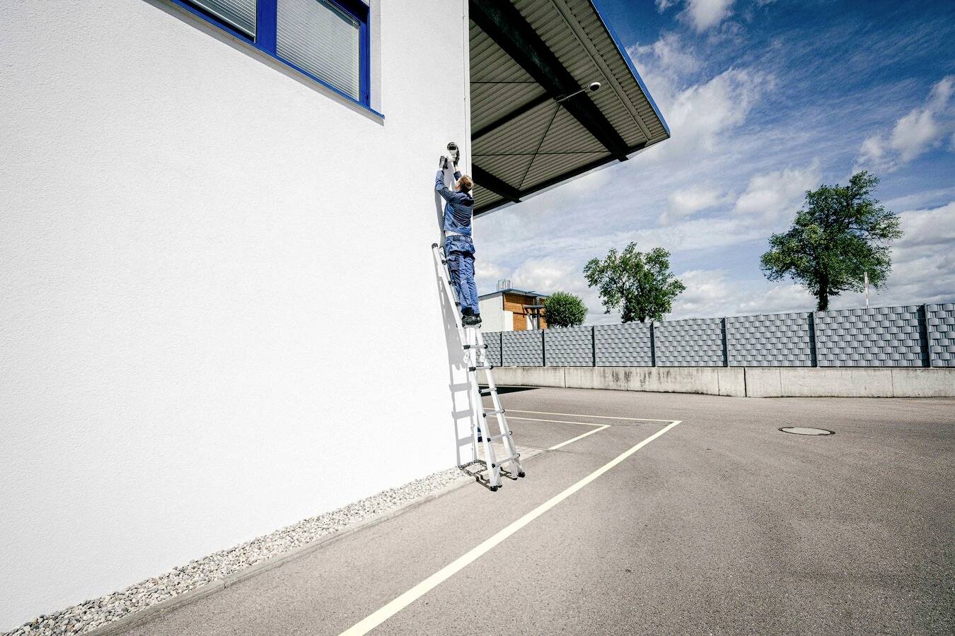 Ein Arbeiter in blauer Uniform steht auf einer Leiter und montiert etwas an einer hohen weißen Wand. Im Hintergrund sind Bäume zu sehen.