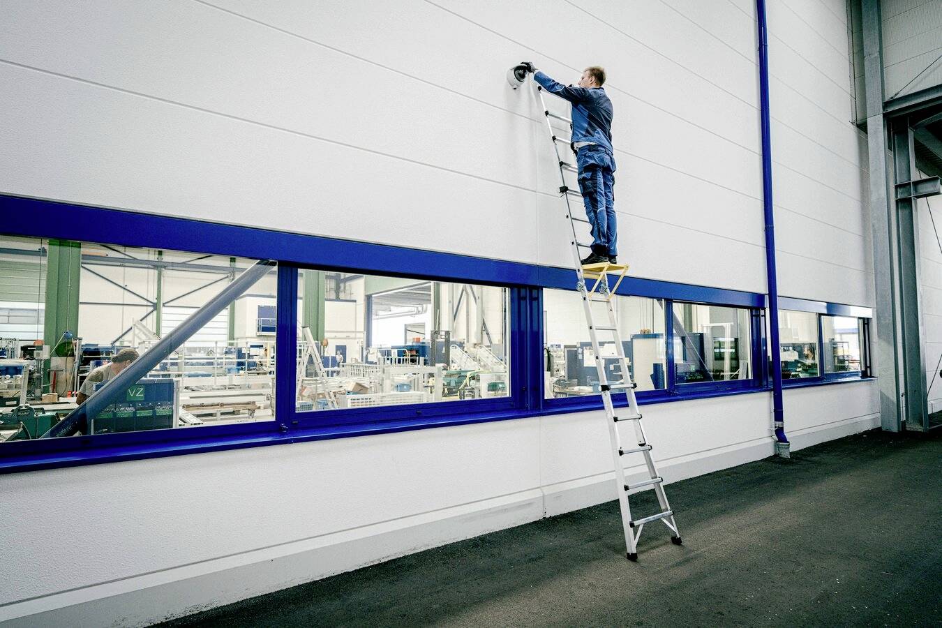 Ein Mann in Arbeitskleidung steht auf einer Leiter und montiert eine Überwachungskamera an der Wand eines Industriegebäudes.