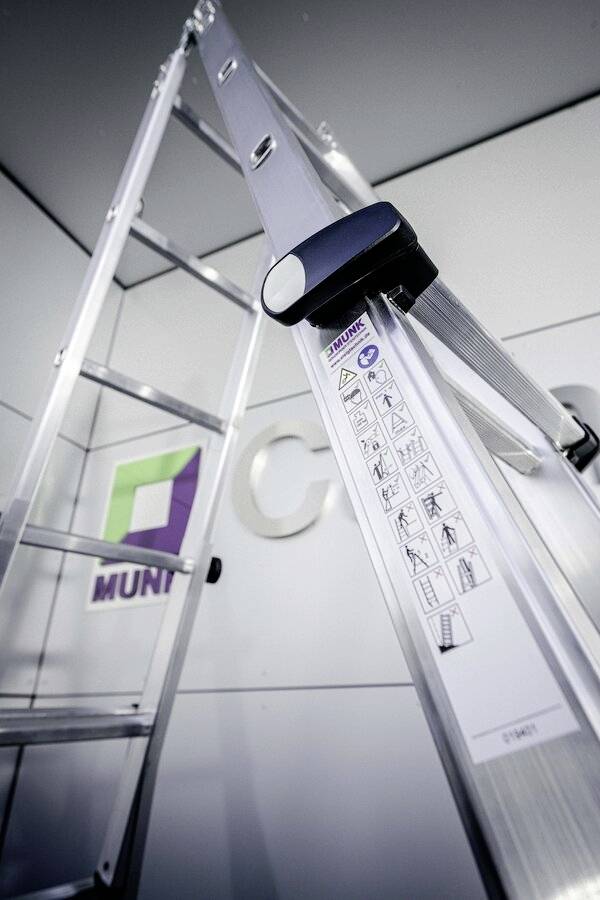 'Aluminiumleiter gegen Wand gelehnt, mit eingezeichneten Sicherheitshinweisen. Firmenlogo im Hintergrund.'