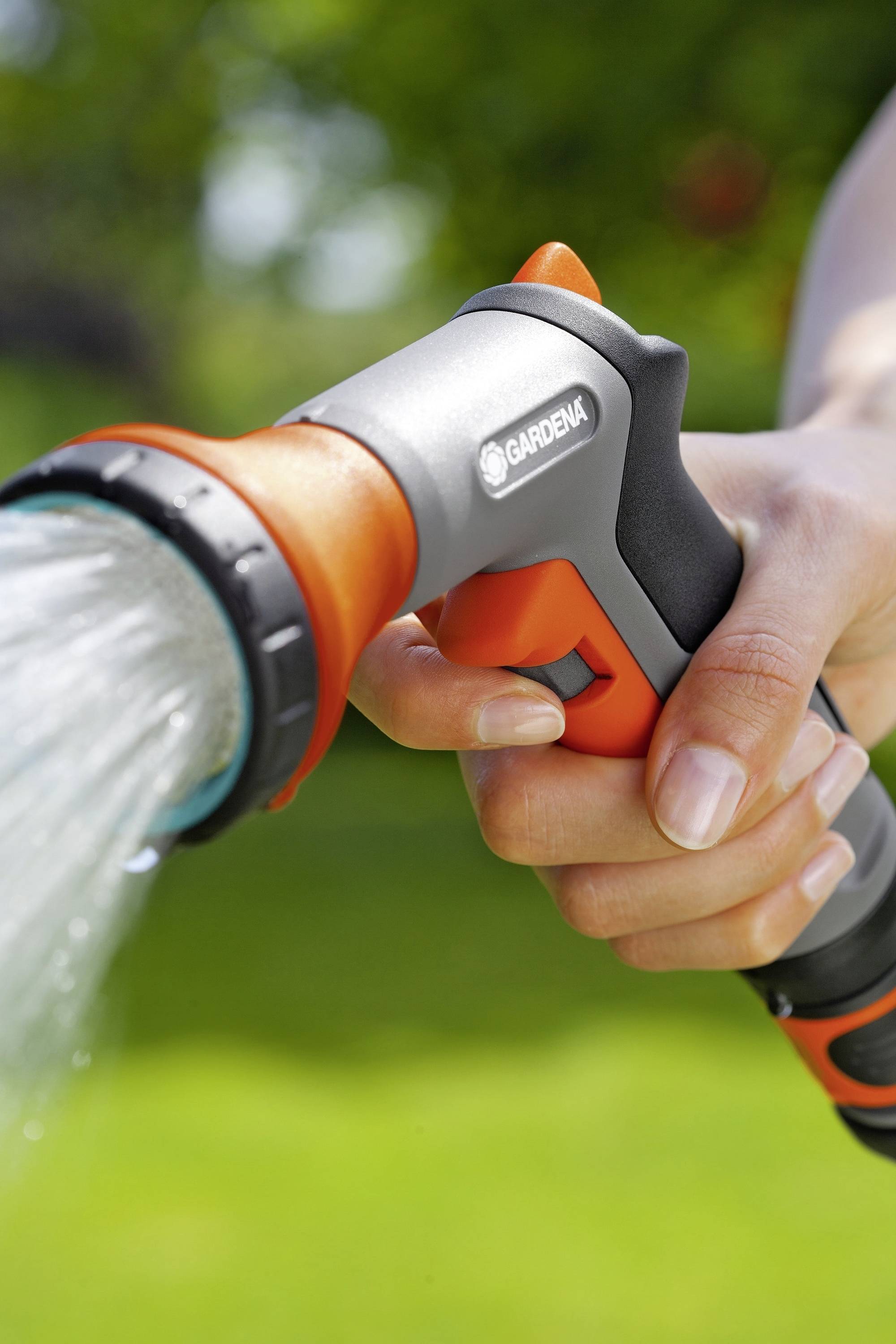 Eine Hand hält eine Gartenbrause, aus der Wasser sprüht, im Vordergrund eines grünen Gartens.
