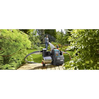 Ein Mann gießt mit einem Gartenschlauch im Garten. Im Vordergrund steht eine elektrische Wasserpumpe auf Holzplanken.