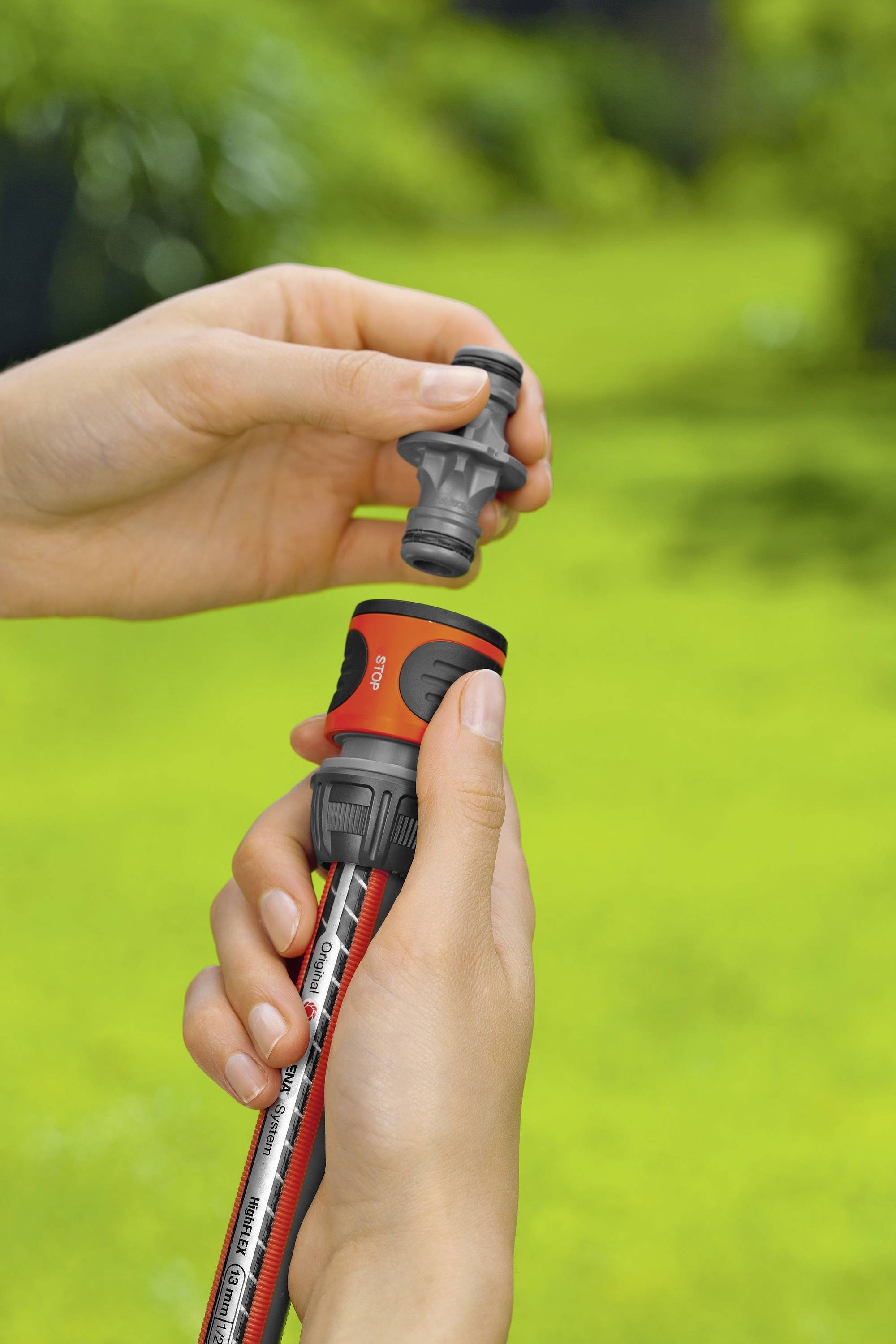 Person hält einen Gartenschlauch mit roter Düse in der Hand und schließt ihn an einen grauen Schlauchverbinder an. Hintergrund unscharf.