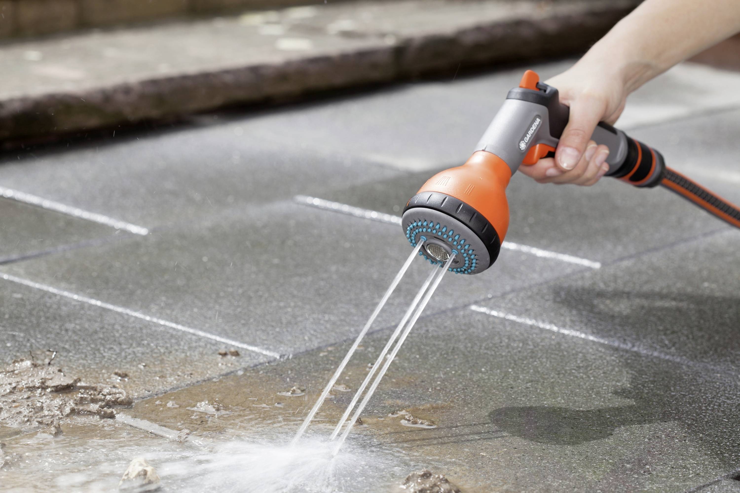 Eine Hand hält eine orangefarbene Sprühdüse, die Wasser in einem Strahl auf den steinernen Boden sprüht.