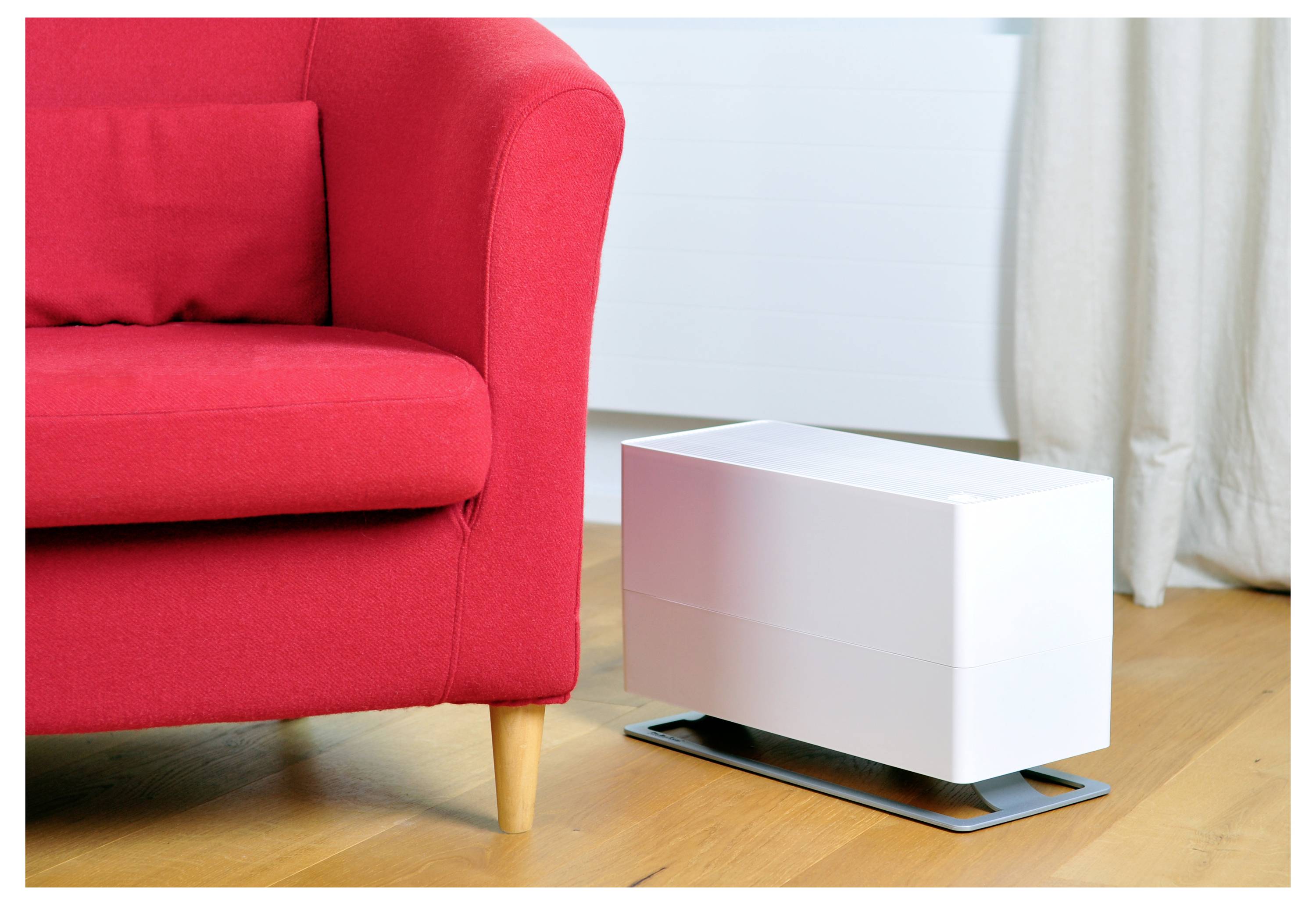 Ein rotes Sofa neben einem weißen Luftreiniger auf einem Holzboden in einem hellen Raum mit Vorhängen.