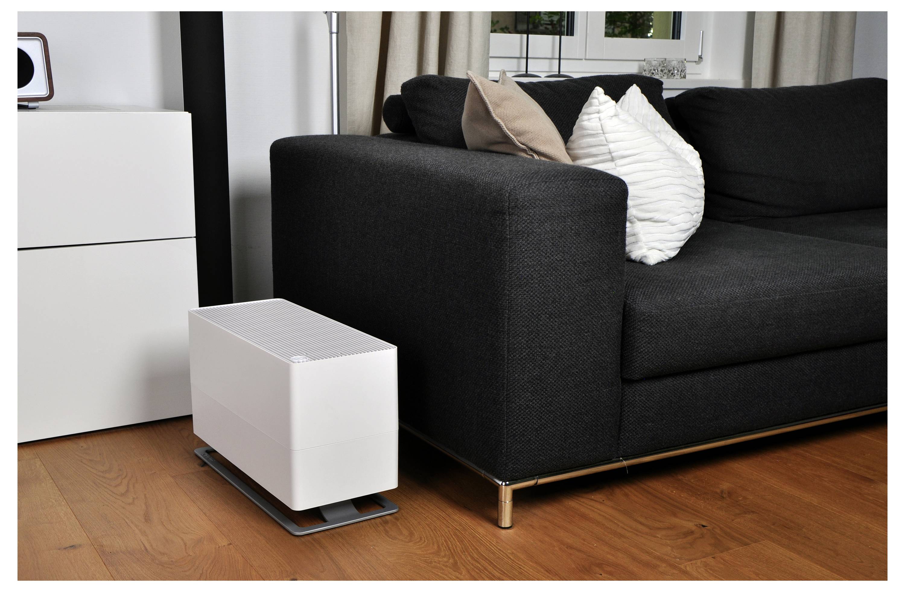Ein weißer tragbarer Luftreiniger steht neben einem schwarzen Sofa in einem modernen Wohnzimmer mit Holzböden.