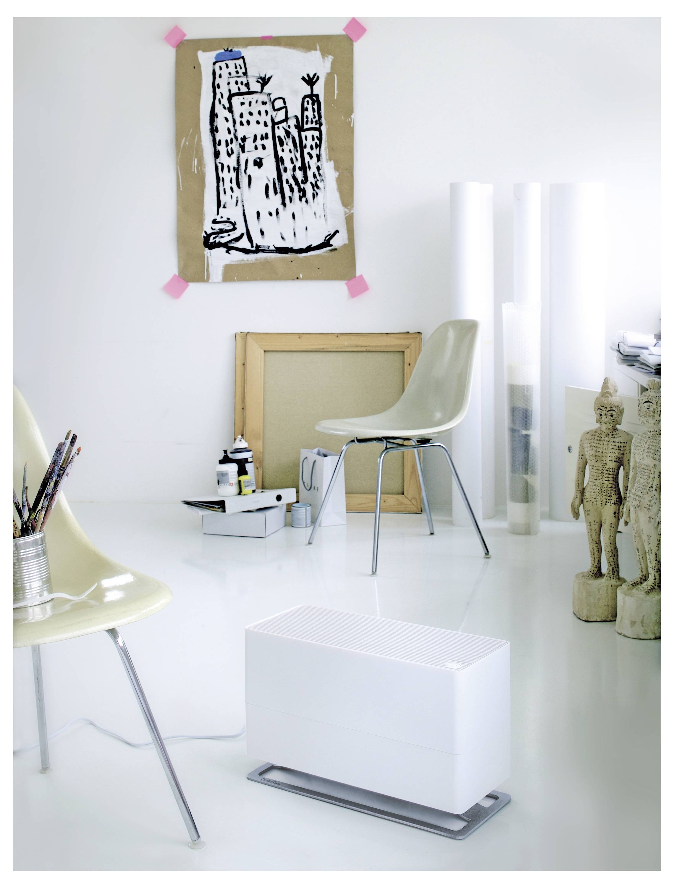 Minimalistische Kunstwerkstatt mit Stühlen, Skizzen und gestapelten Kunstmaterialien; eine mit Klebeband befestigte Stadtzeichnung hängt an der Wand neben einer Skulptur.