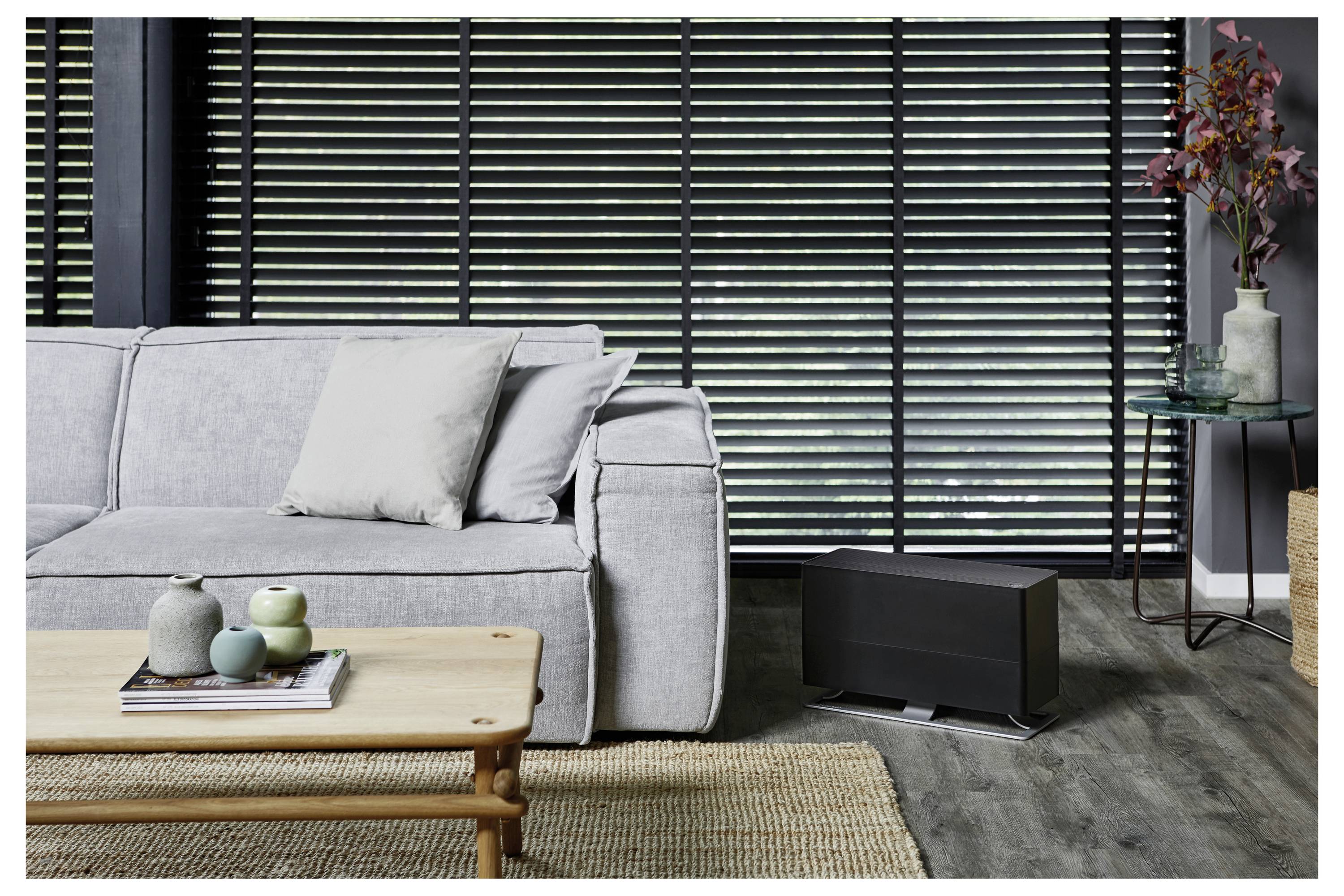 Modernes Wohnzimmer mit grauem Sofa, hölzernem Couchtisch, schwarzem Lowboard und Beistelltisch mit Blumen vor dunklen horizontalen Jalousien.