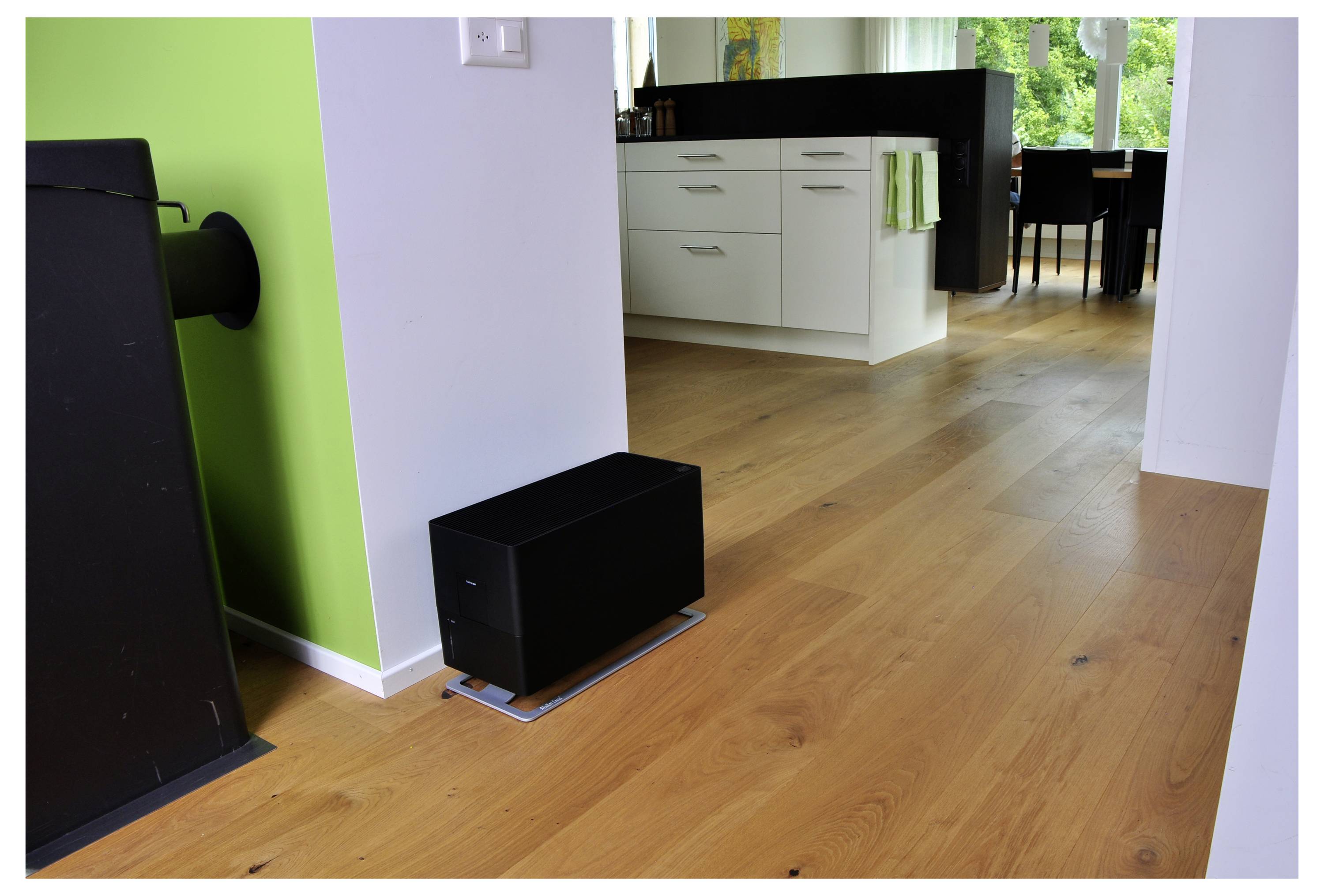 Moderne Küchen- und Essbereich mit Holzfußboden, weißen Schränken, grüner Akzentwand und einem schwarzen Lautsprecher oder Subwoofer auf dem Boden in der Nähe der Wand.