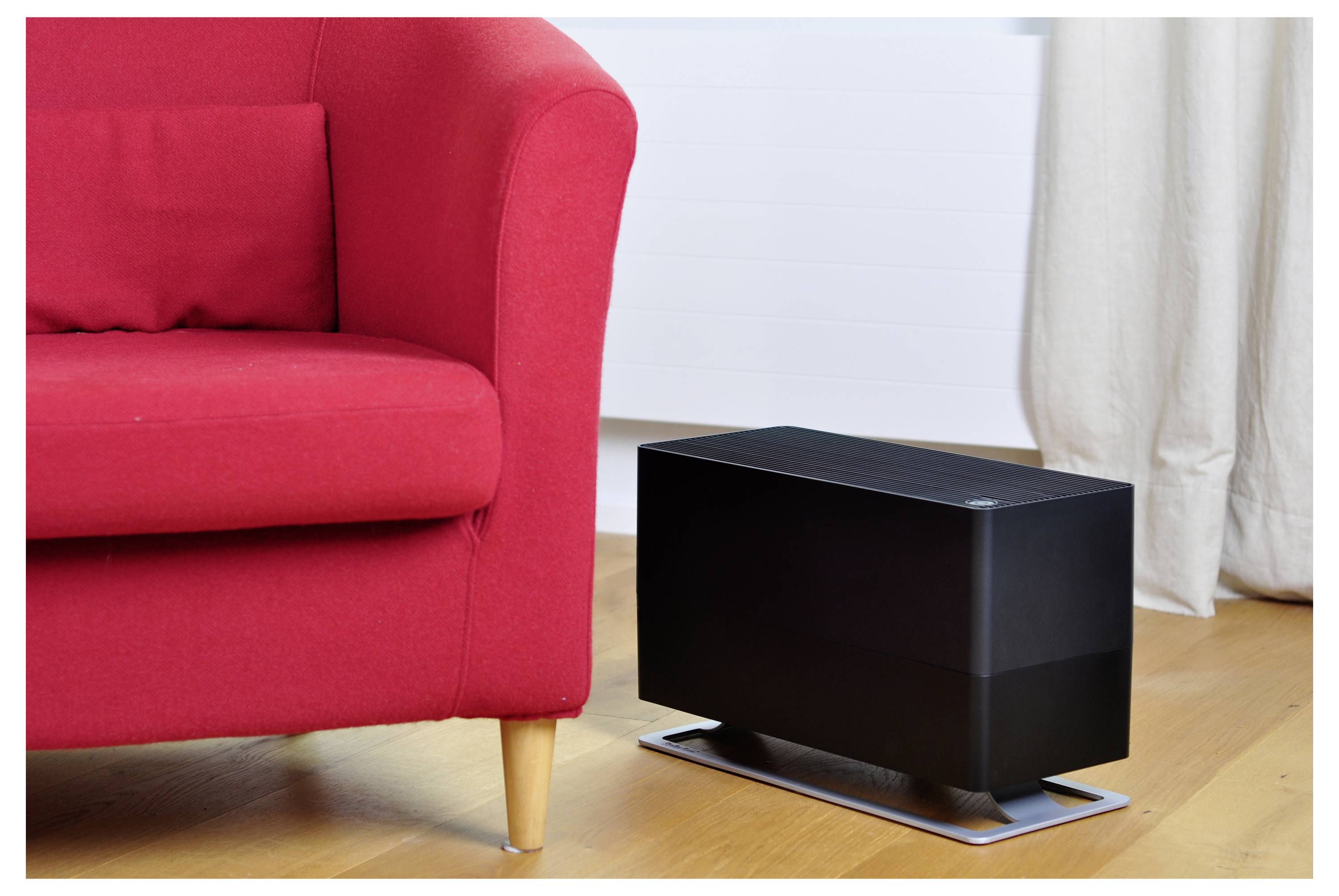 Schwarzer rechteckiger Luftreiniger neben einem roten Sofa auf einem Holzboden in einem hellen Wohnzimmer.