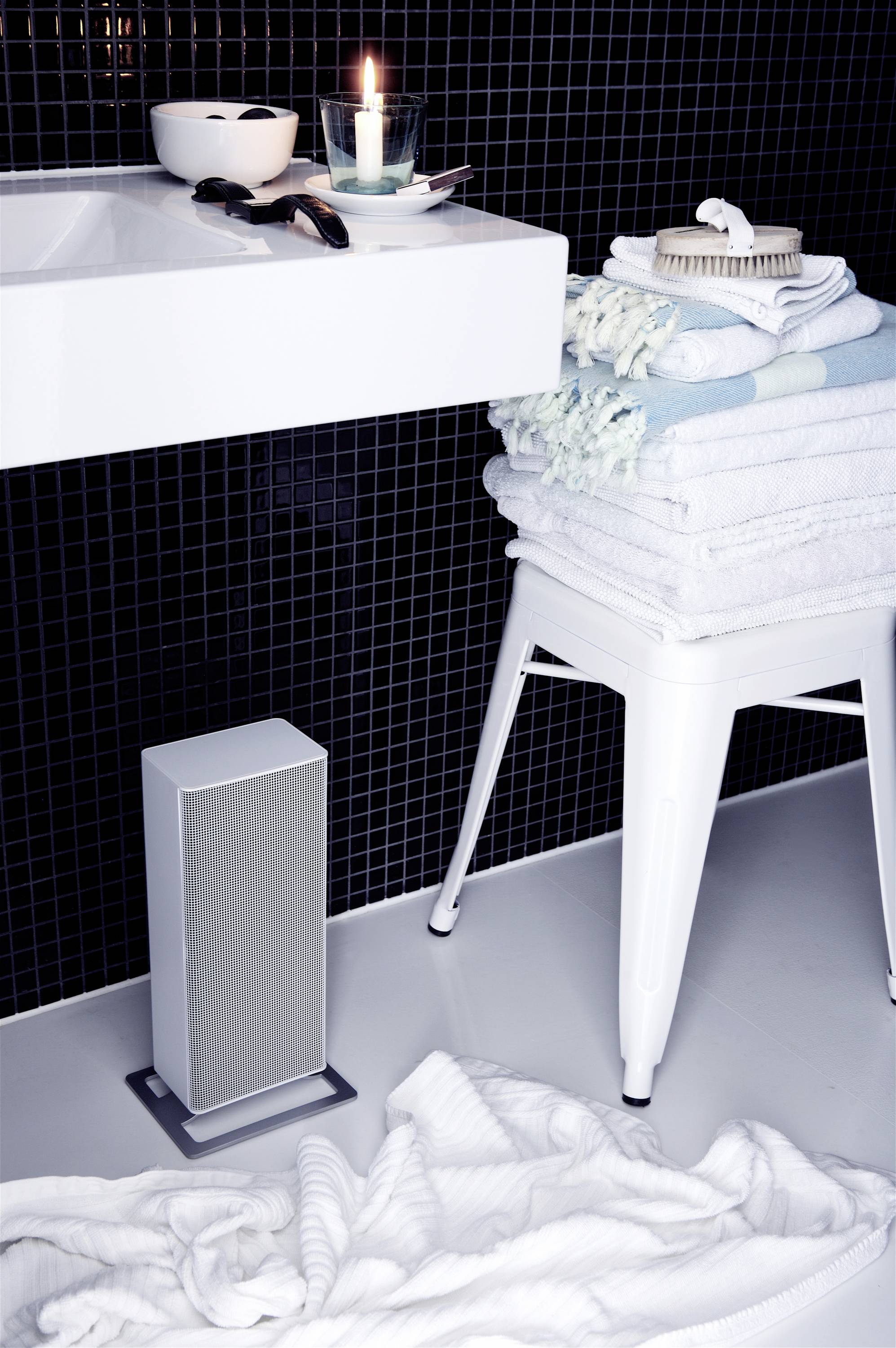 Ein Badezimmer mit einem Waschbecken, einer brennenden Kerze, gefalteten Handtüchern auf einem Hocker und einem Lautsprecher auf dem Boden.