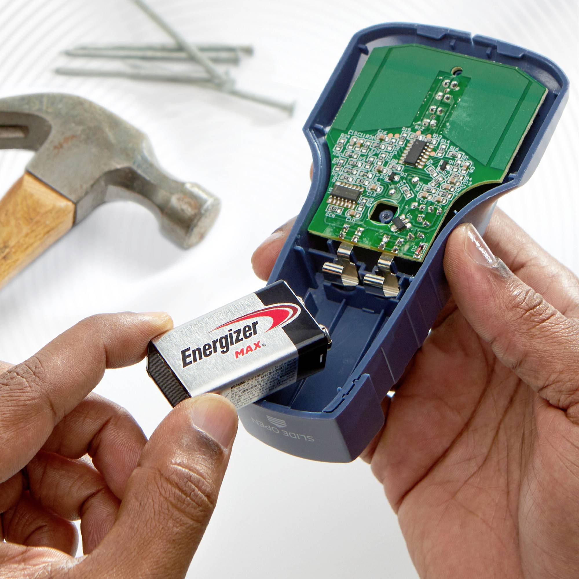 Eine Person hält eine 9-Volt-Batterie neben ein geöffnetes elektronisches Gerät. Im Hintergrund liegt ein Hammer und Nägel.
