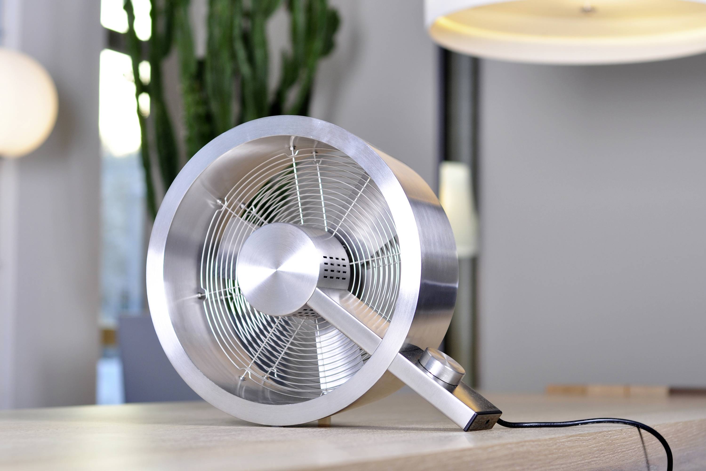 Ein silberner Tischventilator mit einem modernen Design steht auf einem Holztisch. Ein Kaktus und Lampen sind im Hintergrund sichtbar.