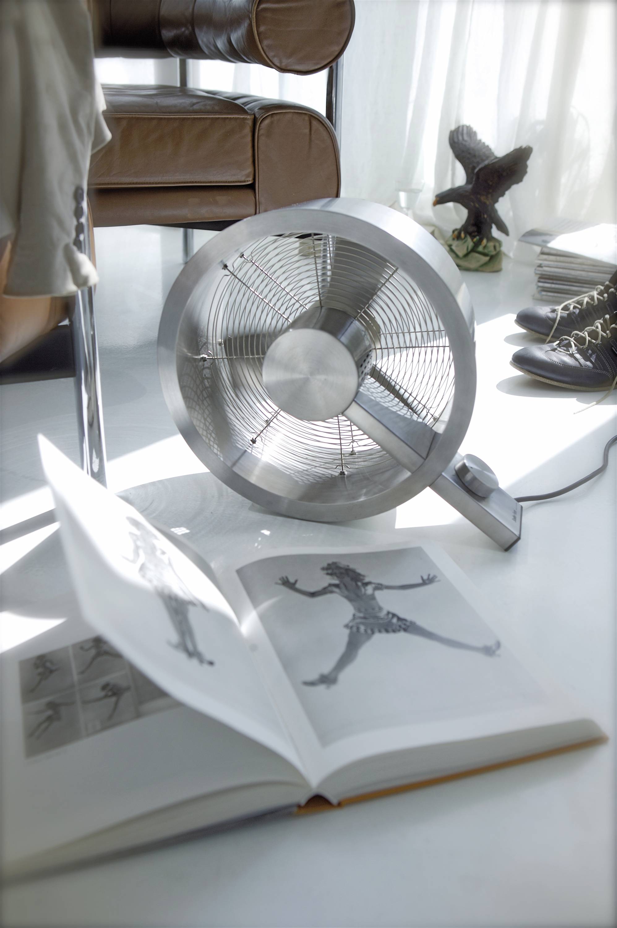 Ein Ventilator steht auf dem Boden neben einem geöffneten Buch mit Schwarz-Weiß-Bildern. Im Hintergrund sind Schuhe und ein Stuhl sichtbar.