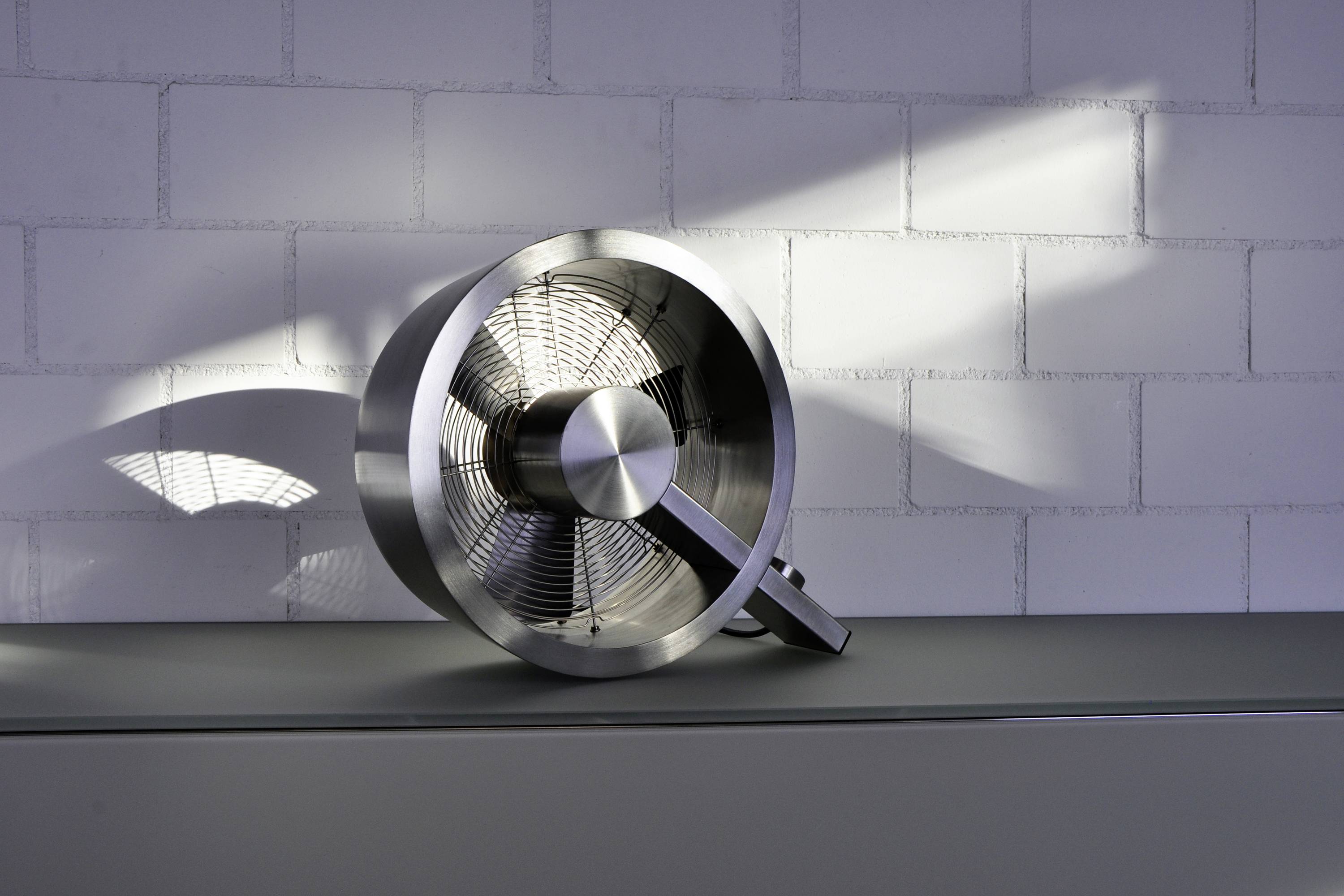 Ein metallischer Ventilator liegt auf der Seite vor einer weißen Backsteinwand. Sonnenlicht wirft Schatten und Licht auf die Wand.